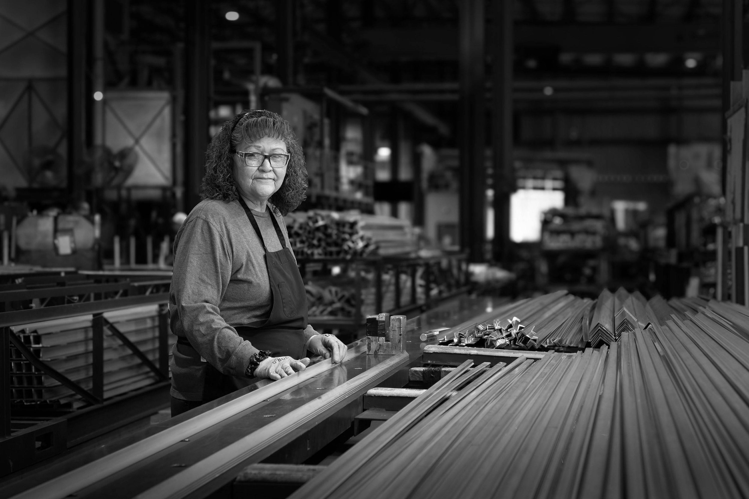 A woman in an apron stands in an industrial factory, working with long aluminum parts on a table, smiling at the camera.