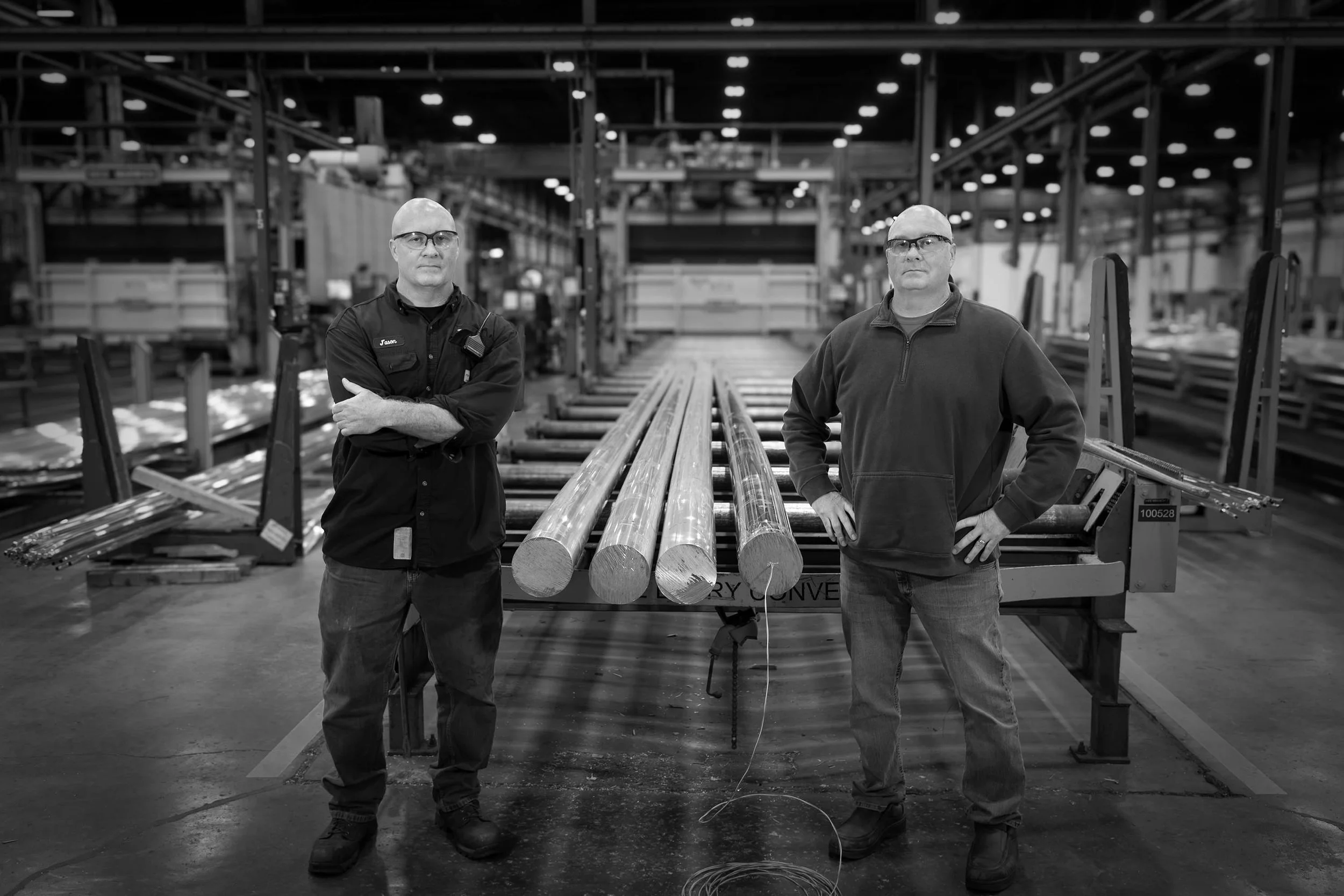 Two men stand in a black and white factory next to long metal rods, with industrial equipment in the background.
