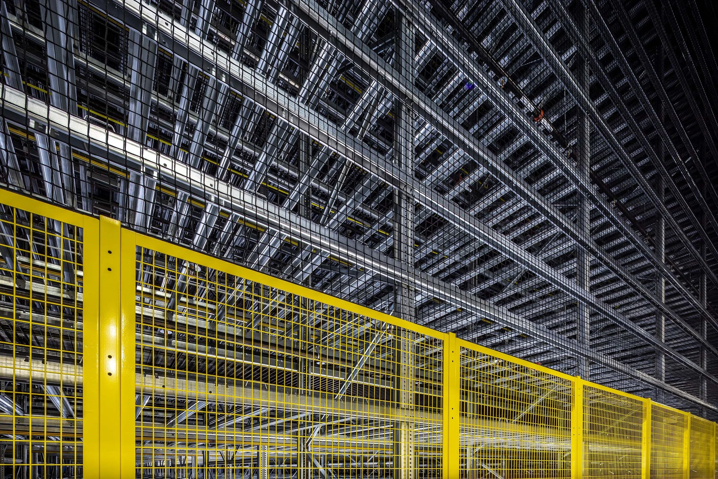 Rows of metal racks and beams form an industrial racking structure behind a yellow wire safety fence, with a complex network of shelves extending into the distance.