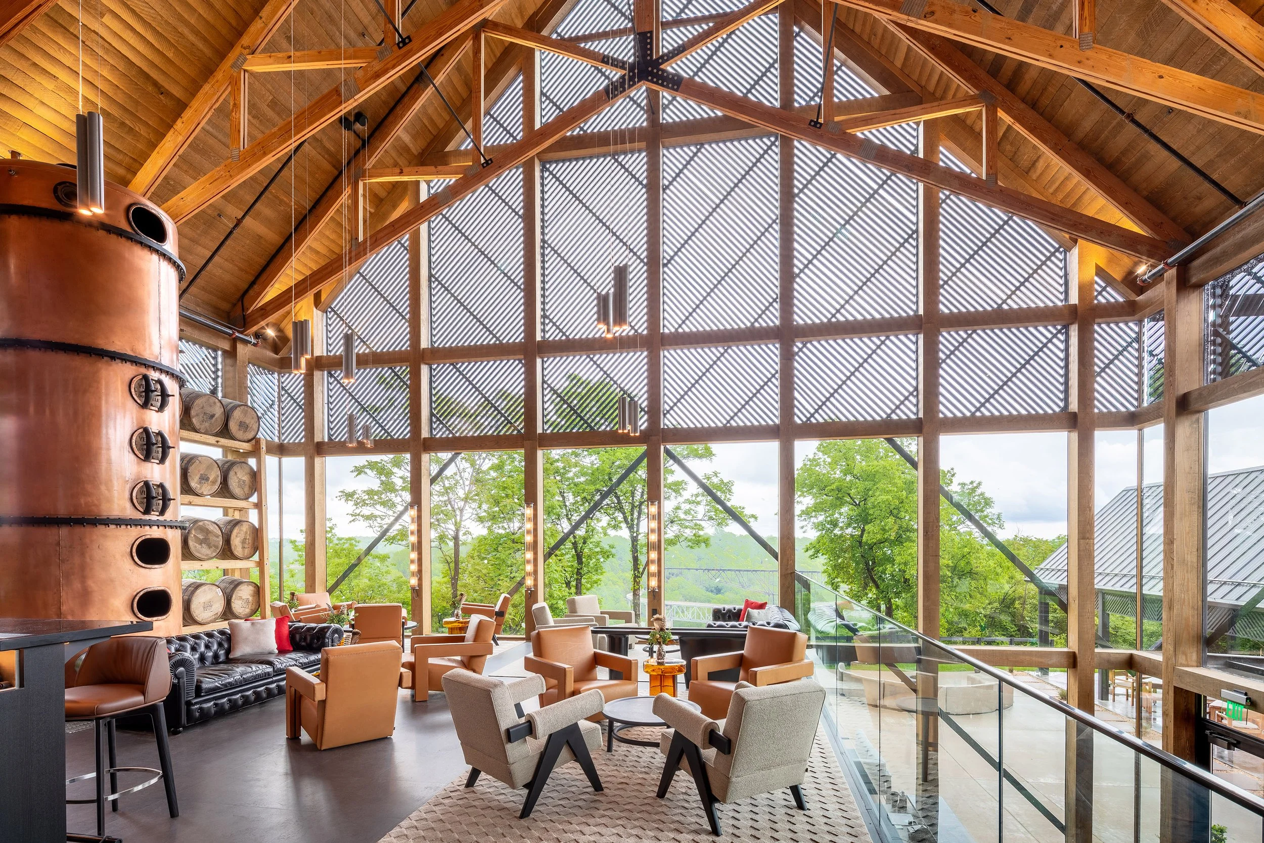 A spacious, modern lounge with wooden beams, floor-to-ceiling windows, tan chairs, and a large copper still reflects striking distillery architecture. Stacked barrels and lush green trees are visible through the glass wall.