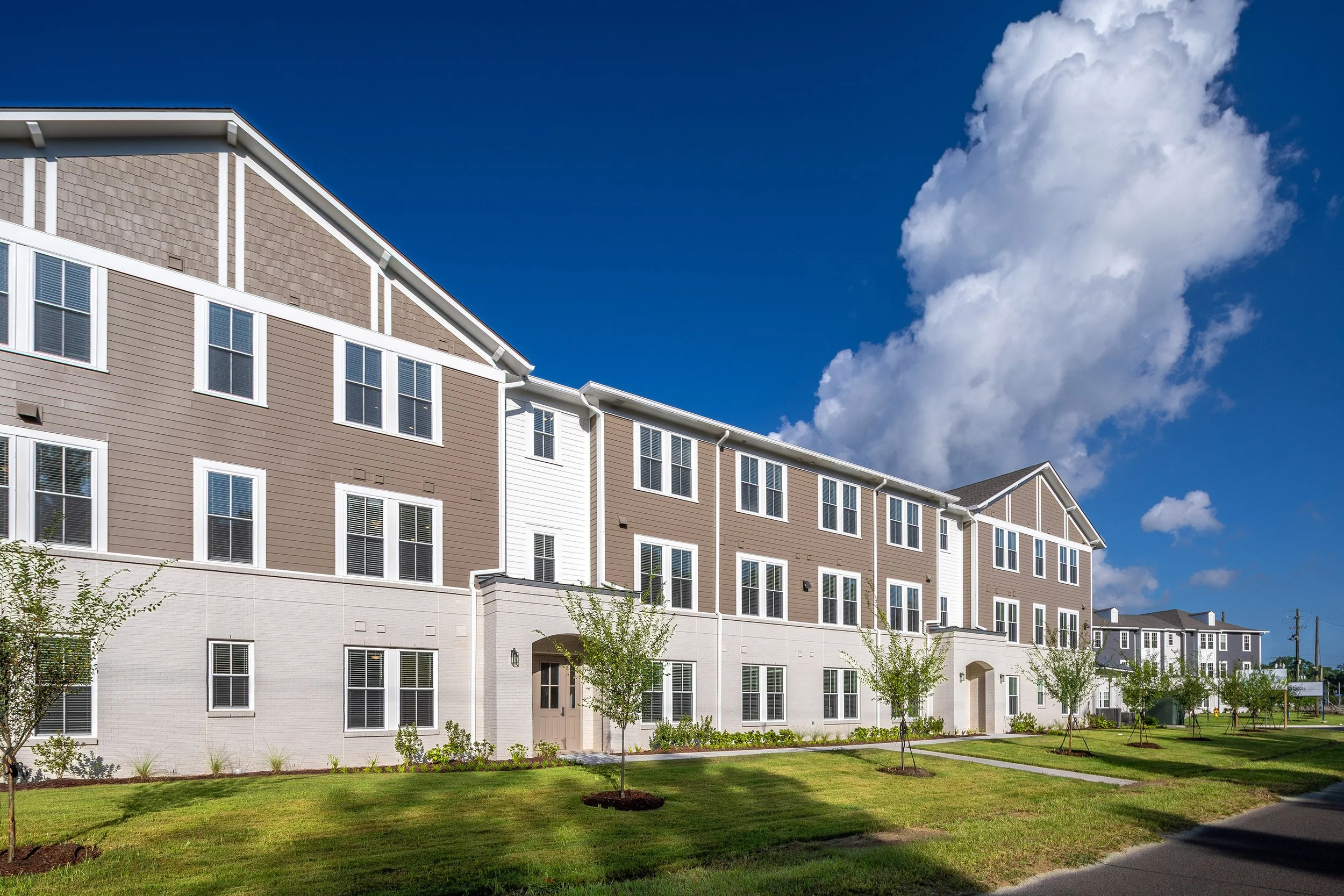 Modern three-story apartment building with beige and white exterior, multiple windows, and a landscaped lawn under a bright blue sky with large, puffy clouds.