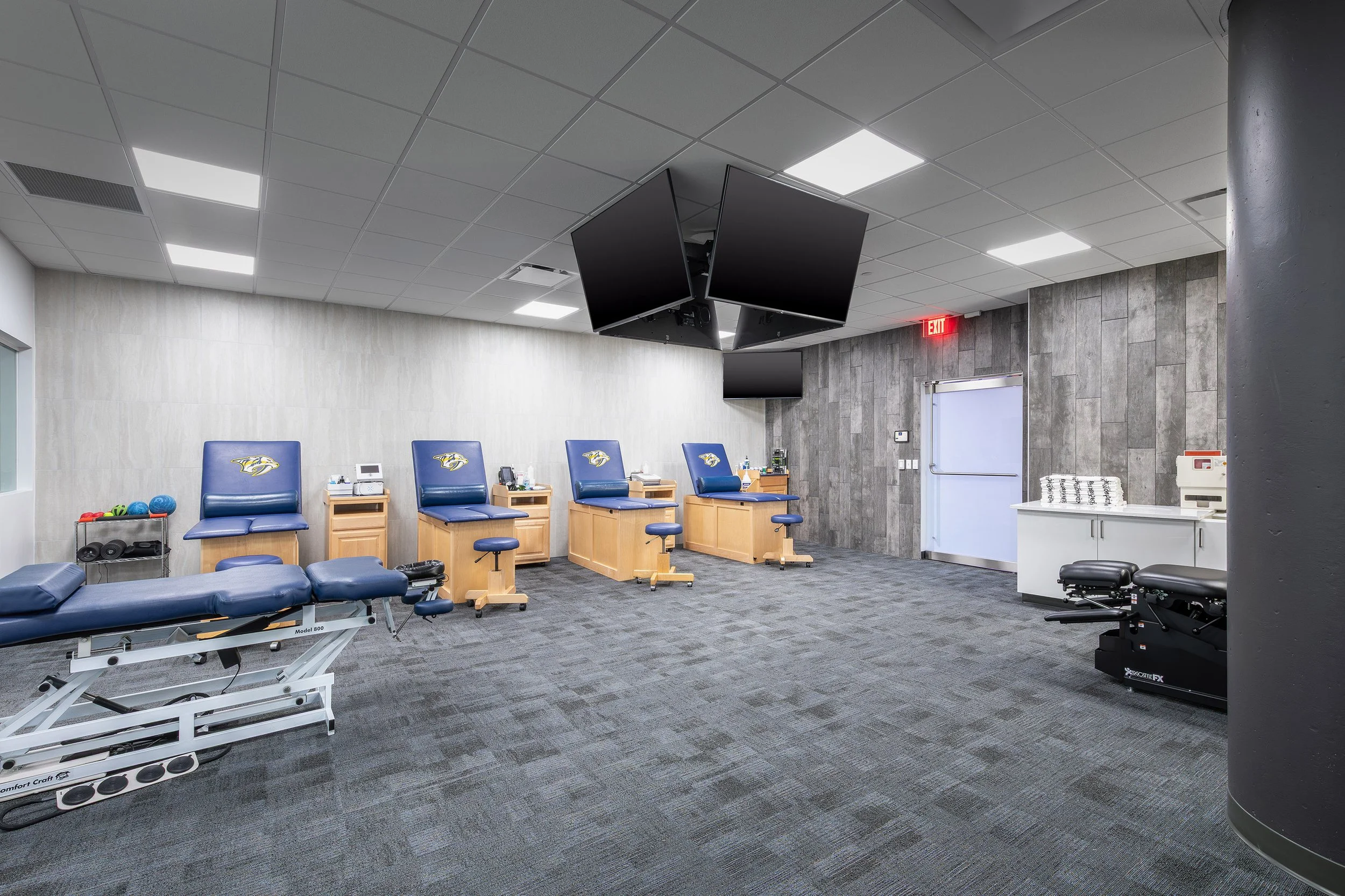 A modern physical therapy room with blue padded tables, wooden cabinets, exercise weights, stacked towels, and wall-mounted TVs playing Predators hockey highlights. The space features bright lighting, gray carpet, and wood-paneled walls.