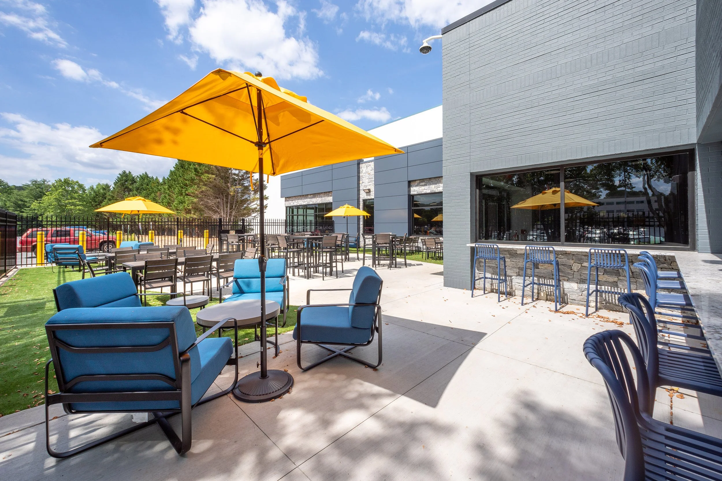 Outdoor employee dining area with blue cushioned chairs, yellow umbrellas, bar stools, and tables next to a modern office building. Trees and a fence are in the background under a partly cloudy sky.