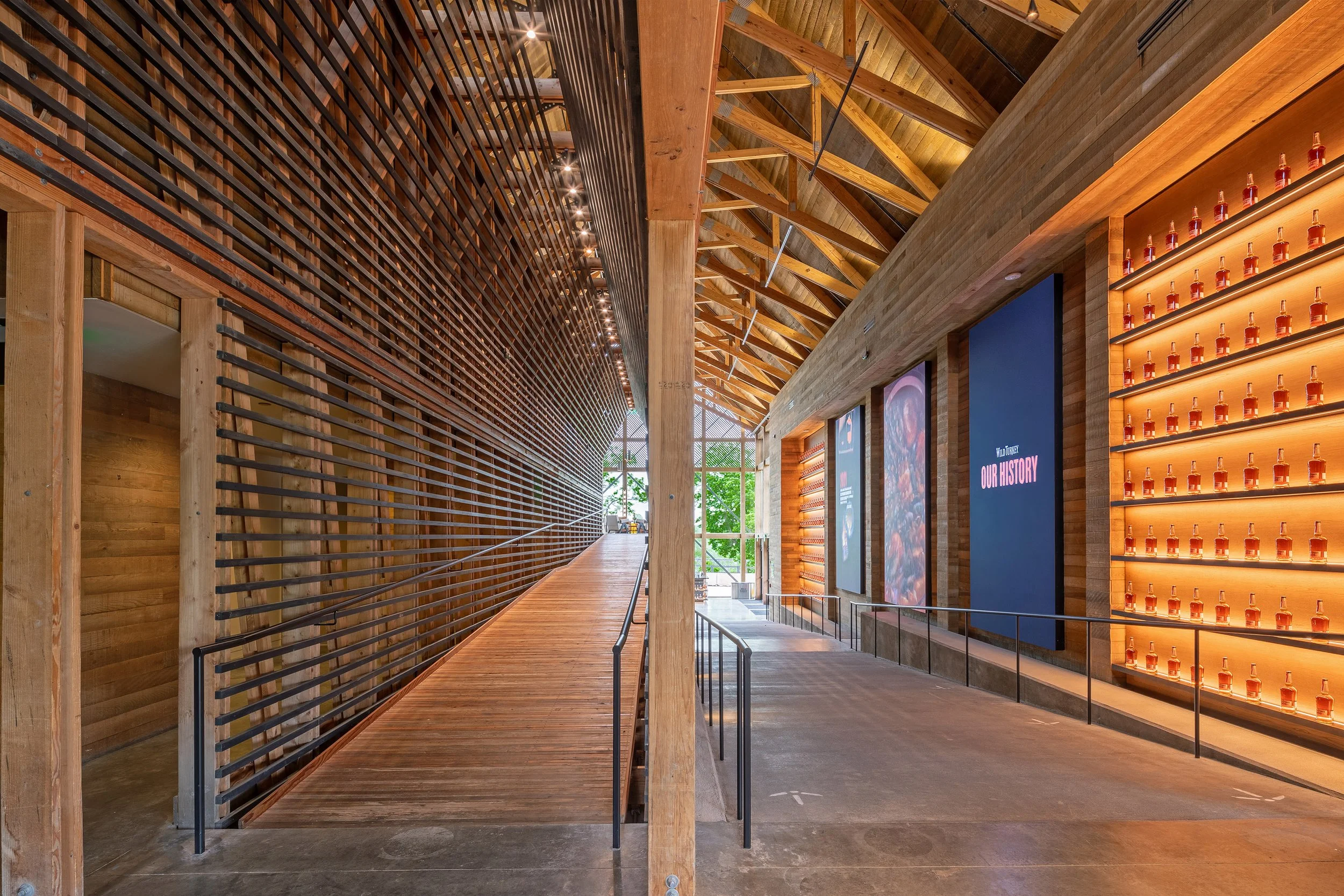 A modern interior with a wooden ramp on the left, tall ceilings with exposed beams, and a wall of shelves displaying bourbon bottles on the right. Natural light filters in from large windows at the end.