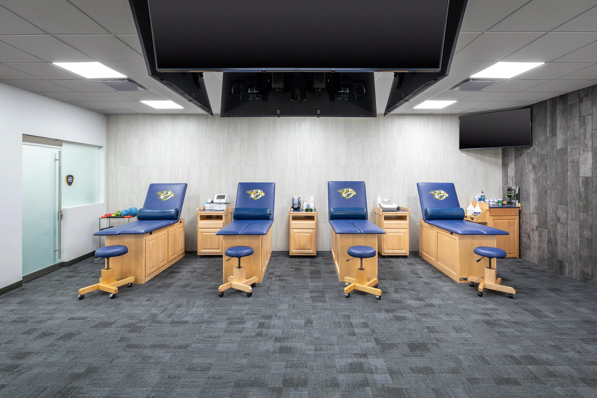 A modern sports medicene treatment room with four blue examination tables, matching stools, wooden cabinets, medical supplies, and bright overhead lighting—perfect for treating Predators players behind the scenes at Bridgestone Arena. A glass door an