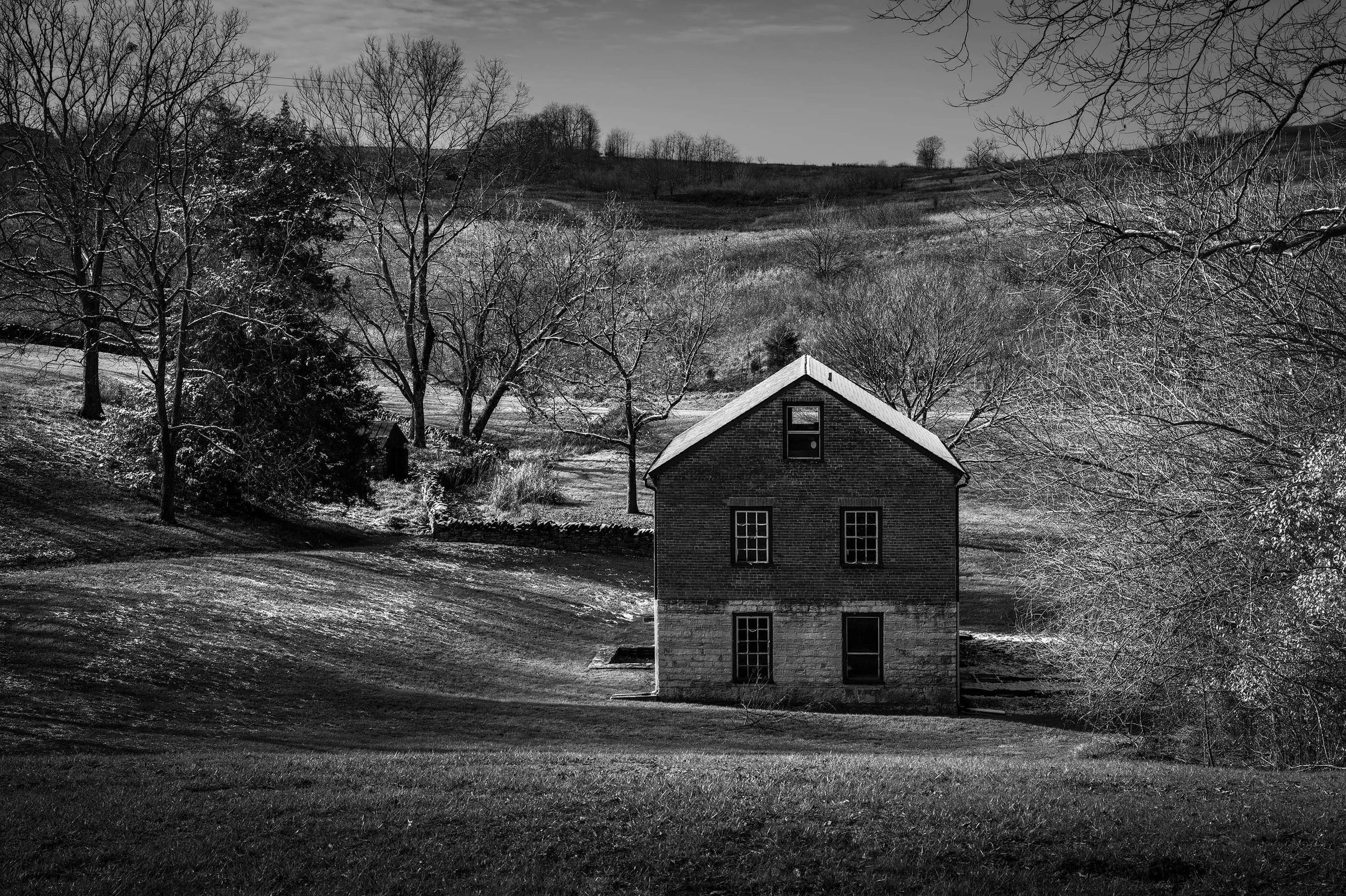 A solitary historic brick house stands in a winter landscape of grassy hills and bare trees, captured in black and white.