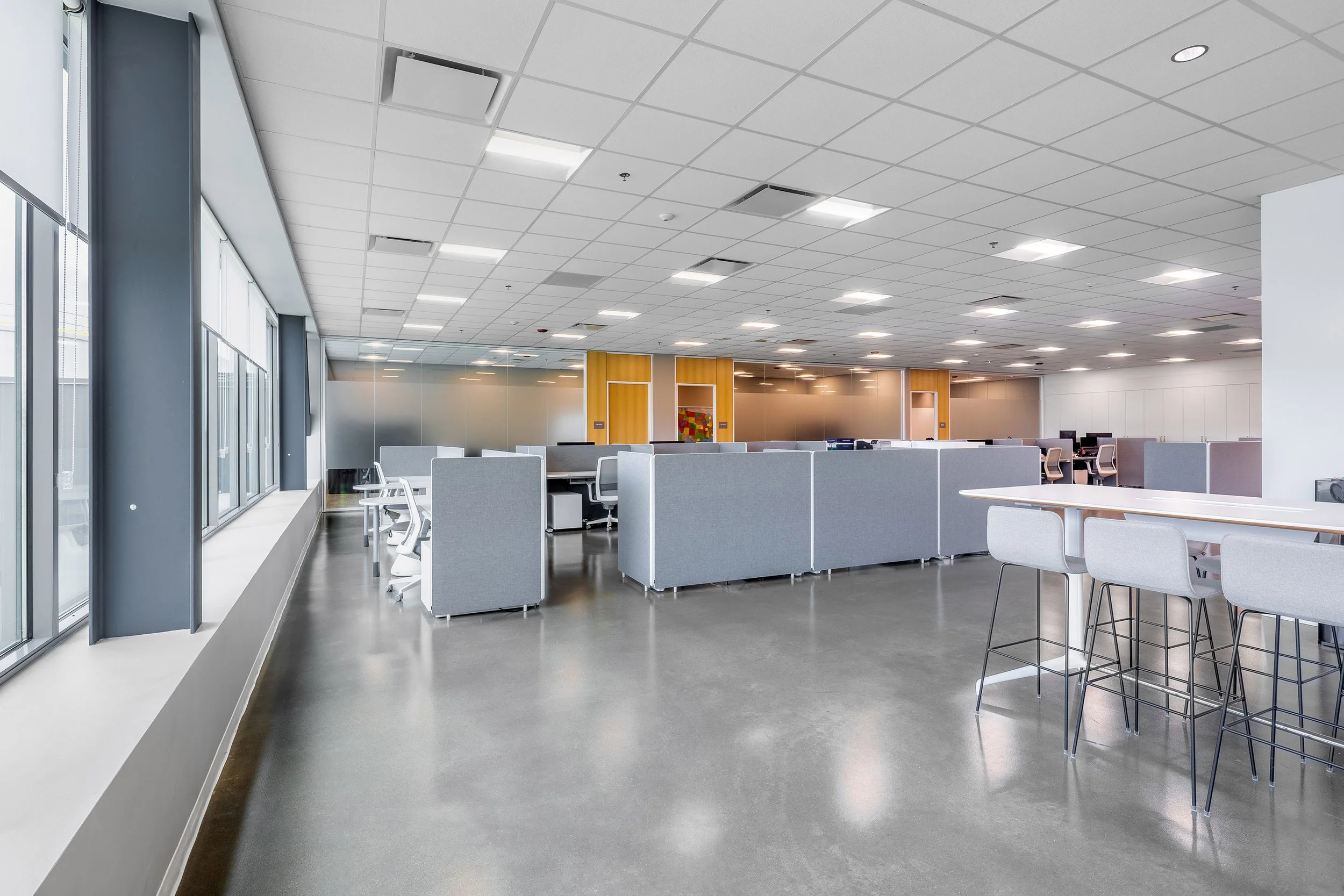 Modern, open-plan office space with gray cubicles, white desks, and office chairs has an industrial feel. High tables with stools and polished concrete floors complement the large windows that fill this architectural area with natural light. Clean an