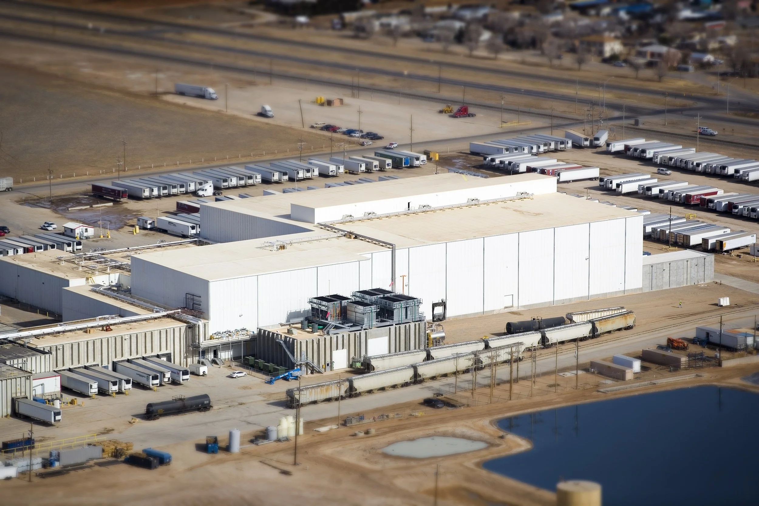 A large industrial warehouse with trucks and trailers parked around it in an area near a retention pond.