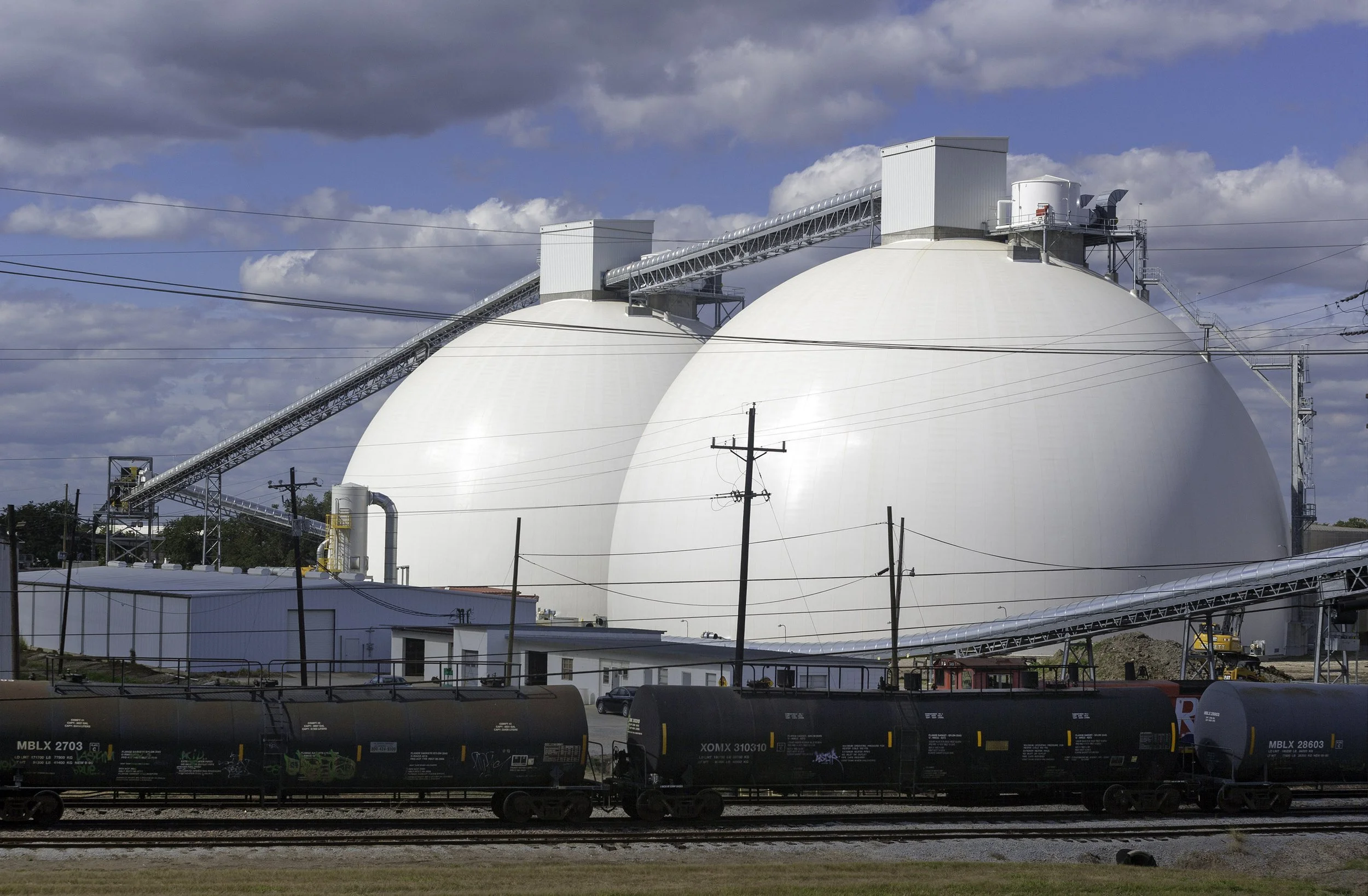 Two large white industrial domes behind train cars and power lines under a partly cloudy sky.