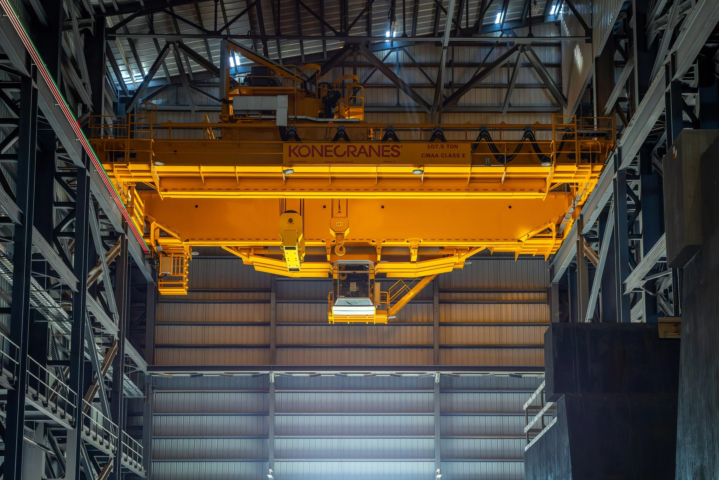 A large yellow overhead crane labeled Konecranes is suspended inside an industrial warehouse with high ceilings and metal beams, capturing the essence of industrial photography.