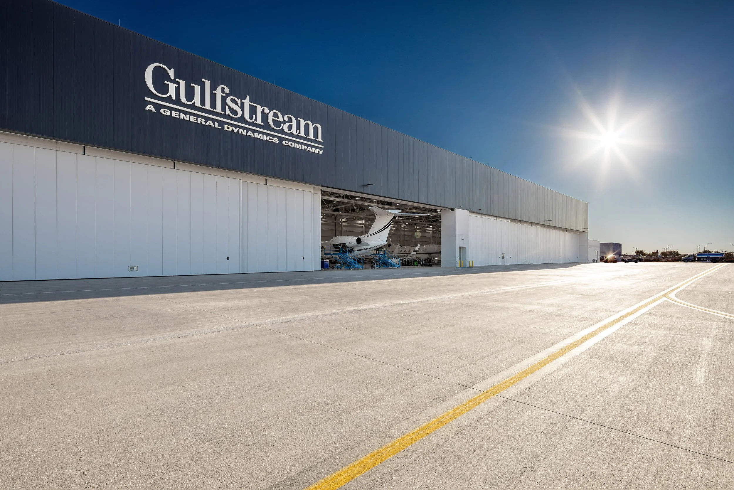 A large Gulfstream hangar with its doors open, revealing private jets inside. The sun shines in a clear blue sky, casting shadows on the wide concrete pavement—an impressive display of aviation and architectural photography.