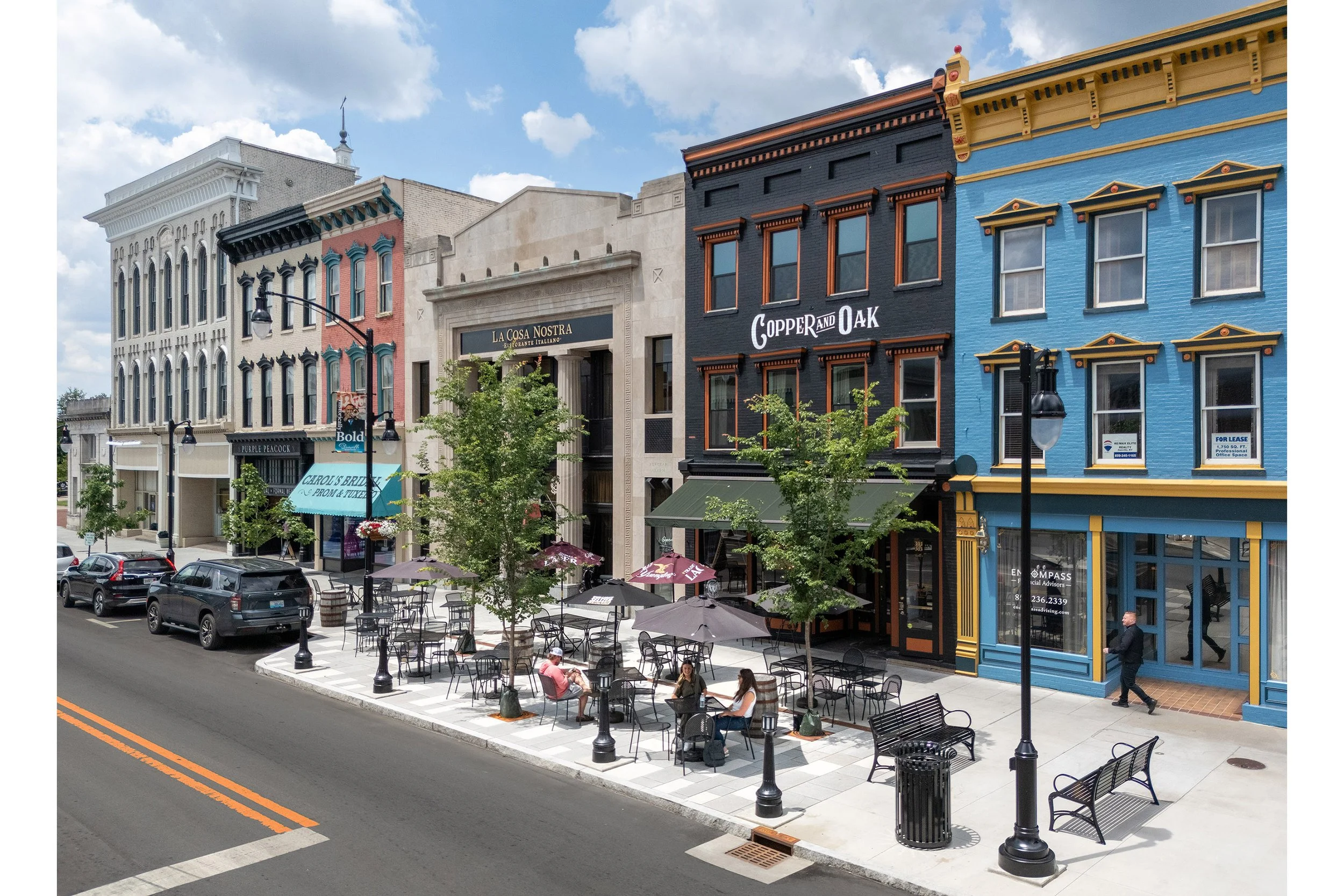 A row of colorful storefronts and restaurants with outdoor seating showcases civic design along a lively main street lined with trees.