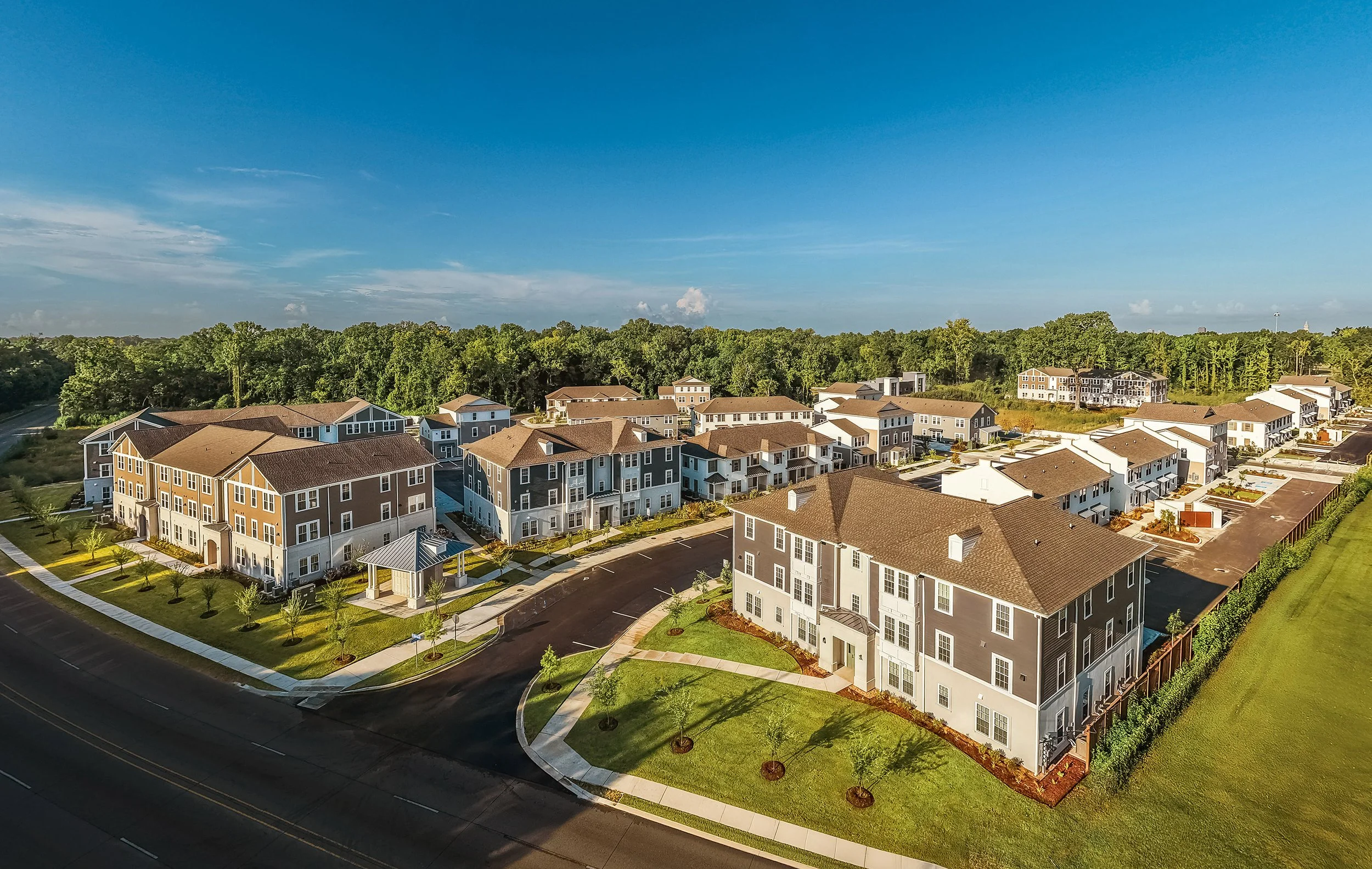 Aerial view of a modern suburban apartment complex with multiple gray and beige buildings, parking lots, green lawns, and surrounding trees under a clear blue sky.
