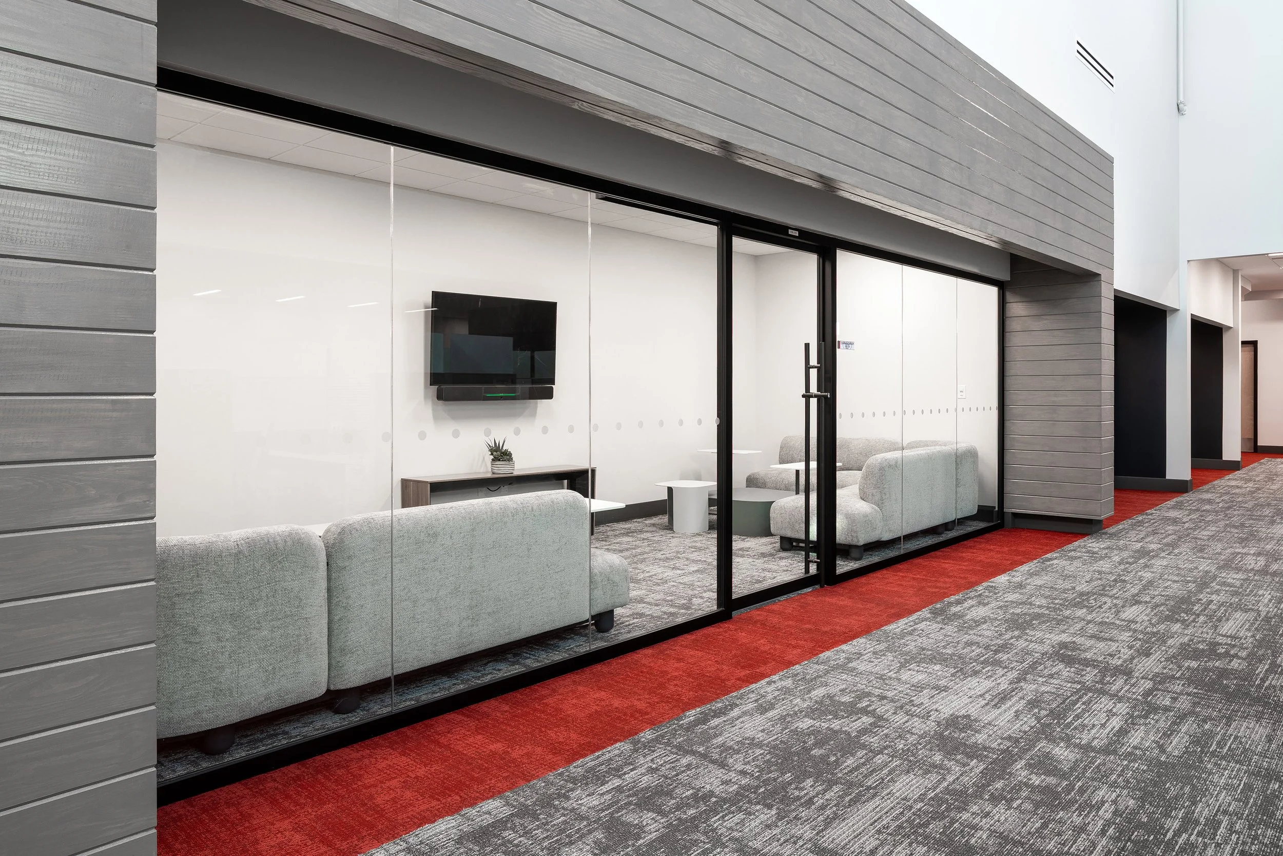 A modern office lounge with glass walls, light gray sofas, a small table, a TV mounted on the wall, and red and gray carpeted floors in the hallway outside.