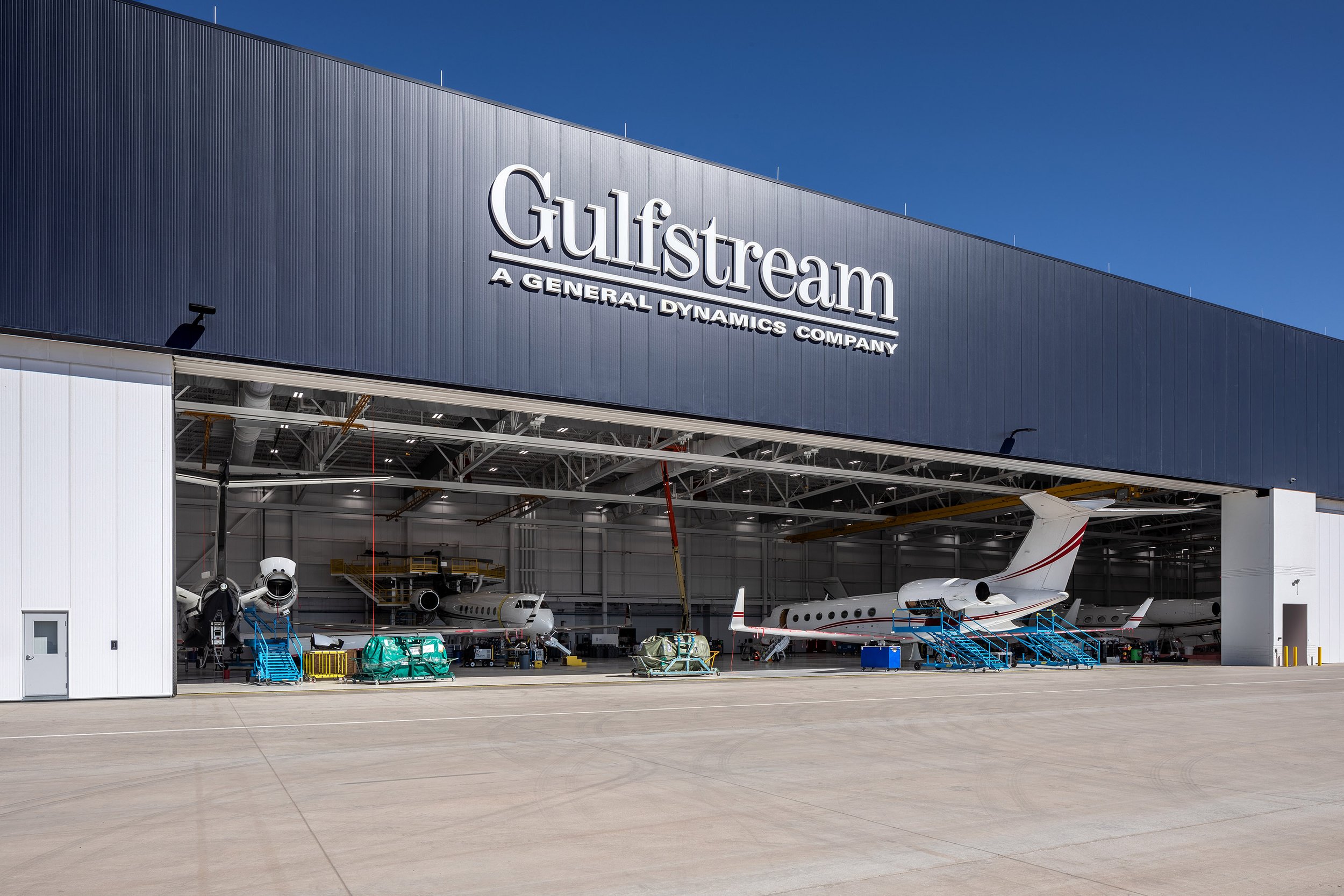 A large Gulfstream aircraft hangar, A General Dynamics Company, showcases aviation photography with multiple private jets under maintenance and various equipment and machinery on the concrete apron outside.