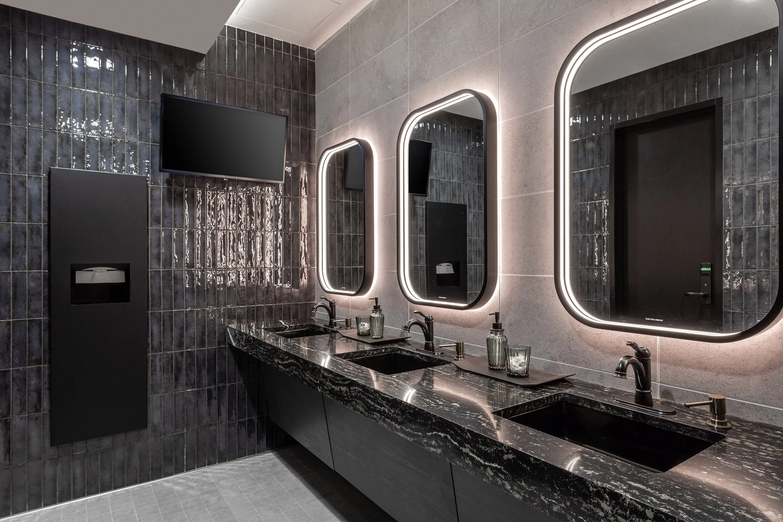 Modern private restroom in the Bridgestone 114 Club Suite features three black marble sinks, large backlit mirrors, dark tiled walls, soap dispensers, a paper towel dispenser, and a wall-mounted TV—all in a sleek, contemporary design.