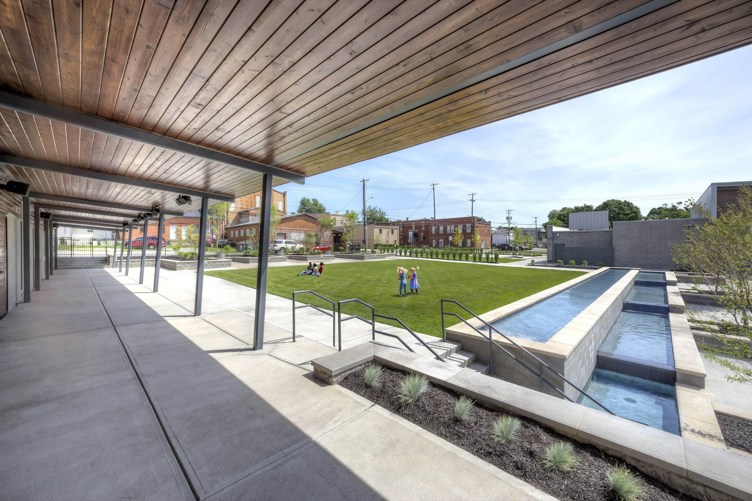 View of a modern outdoor park with a grassy area, children playing, and a water feature, taken from underneath a wooden covered walkway.