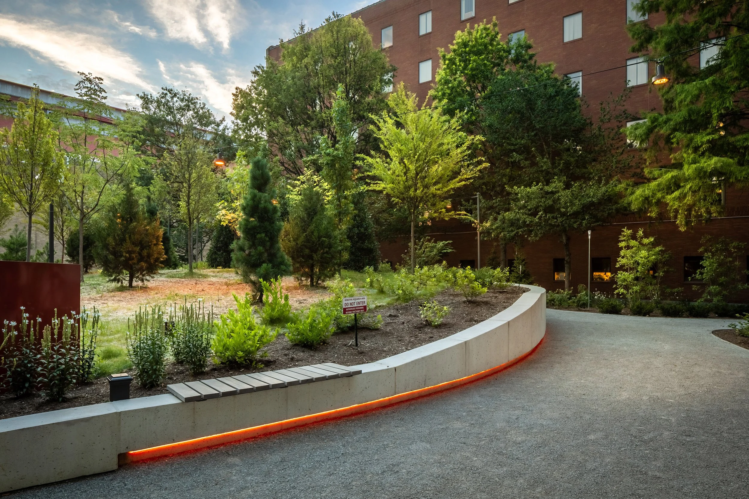 A landscaped urban garden features a curved concrete bench and glowing orange lights, showcasing thoughtful design. Dense trees and plants surround the area, with a large brick building’s architecture in the background under a partly cloudy sky.