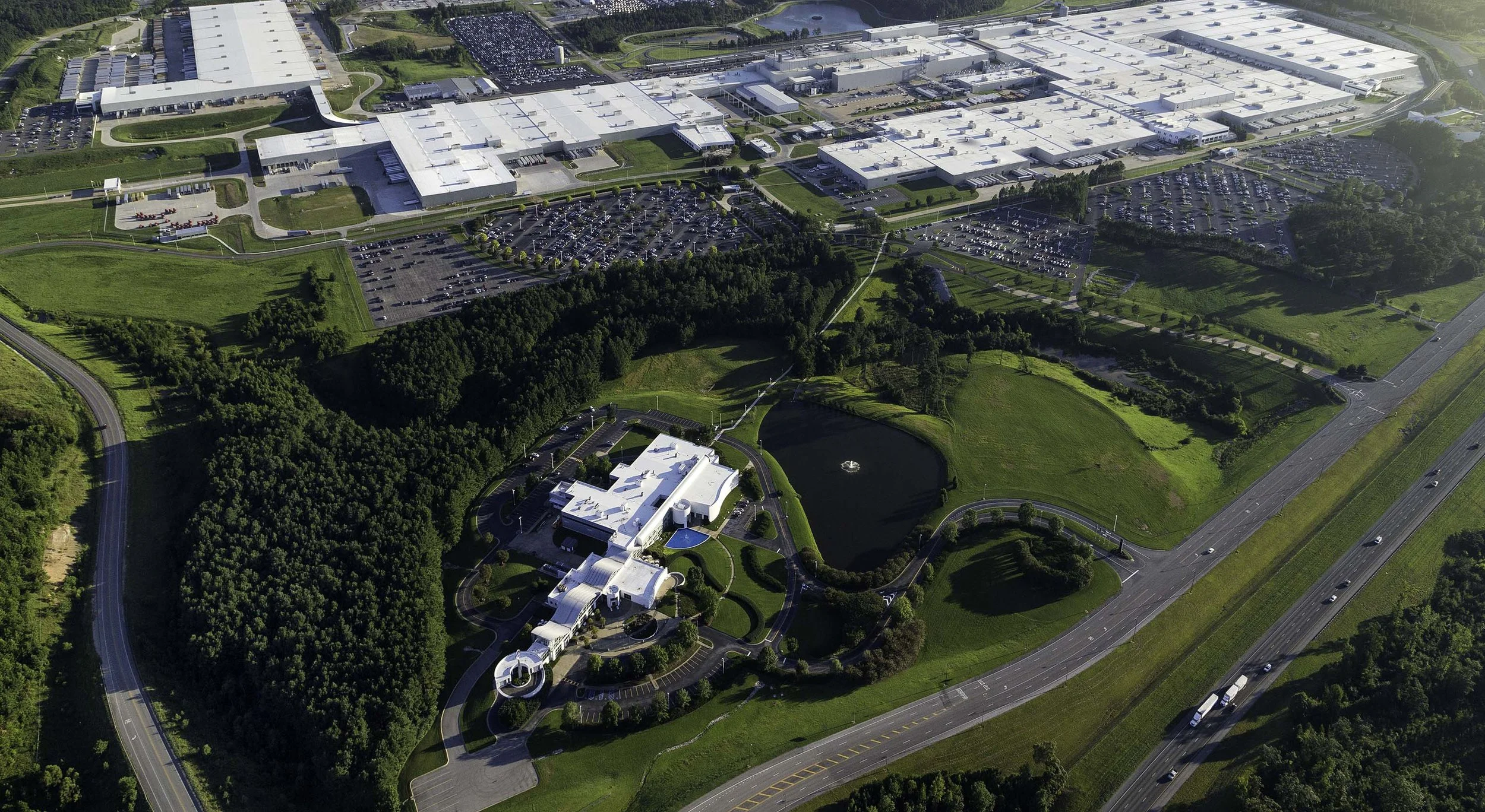 Aerial view of a Mercedes manufacturing facility with white buildings, parking lots, and surrounding green spaces including a pond and wooded areas.