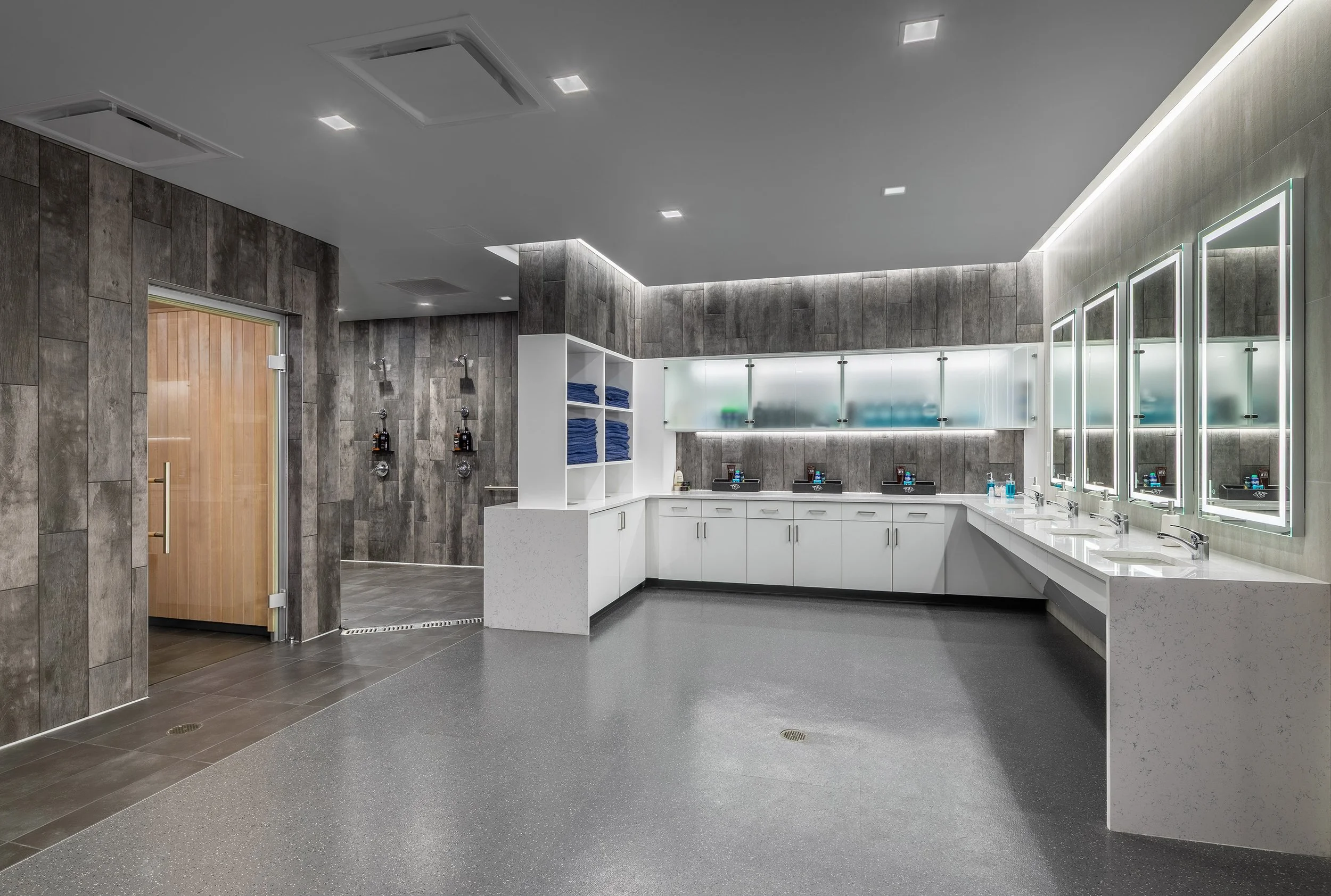 Modern hockey locker room with gray tile walls, white countertops, large mirrors, multiple sinks, open towel storage, and a glass door leading to a sauna. The space is well-lit with recessed and accent lighting—perfect for Predators athletes.