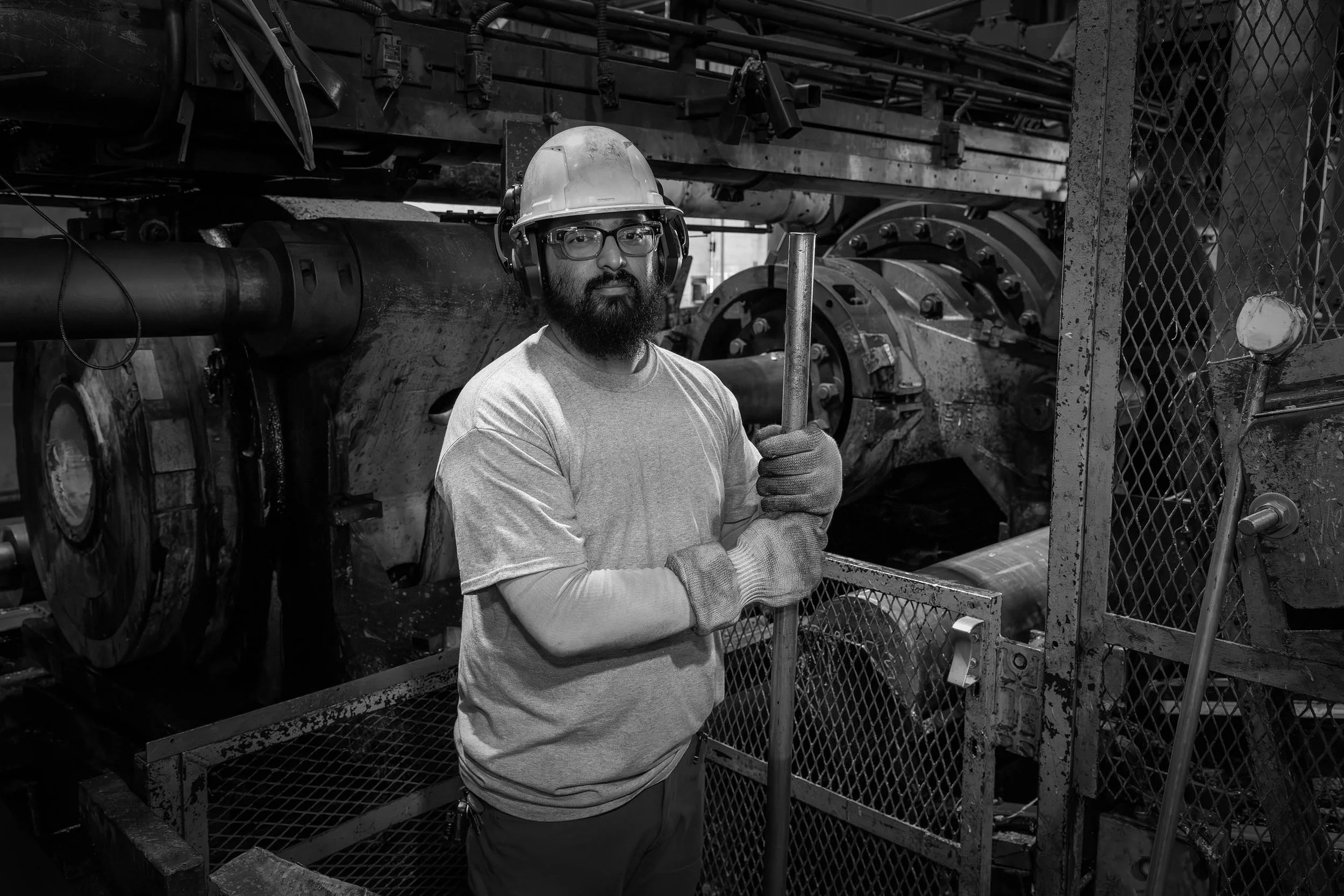 A factory worker in safety gear stands holding an aluminum rod in an industrial setting, looking at the camera.