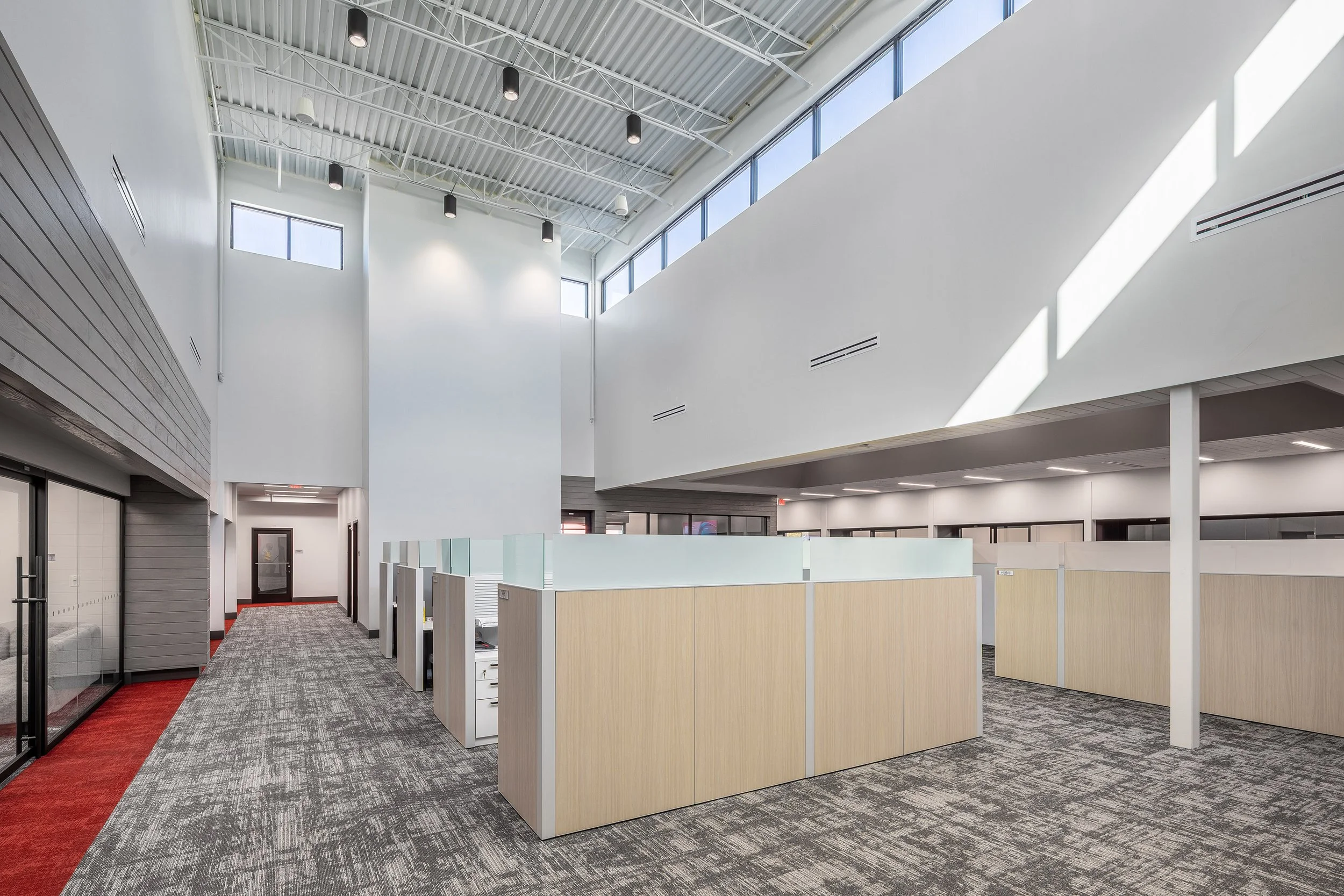 Modern, bright open-plan office with high ceilings, large windows, and rows of cubicles with light wood panels and frosted glass. Red carpet borders the walkway, and gray patterned carpet covers the main floor.