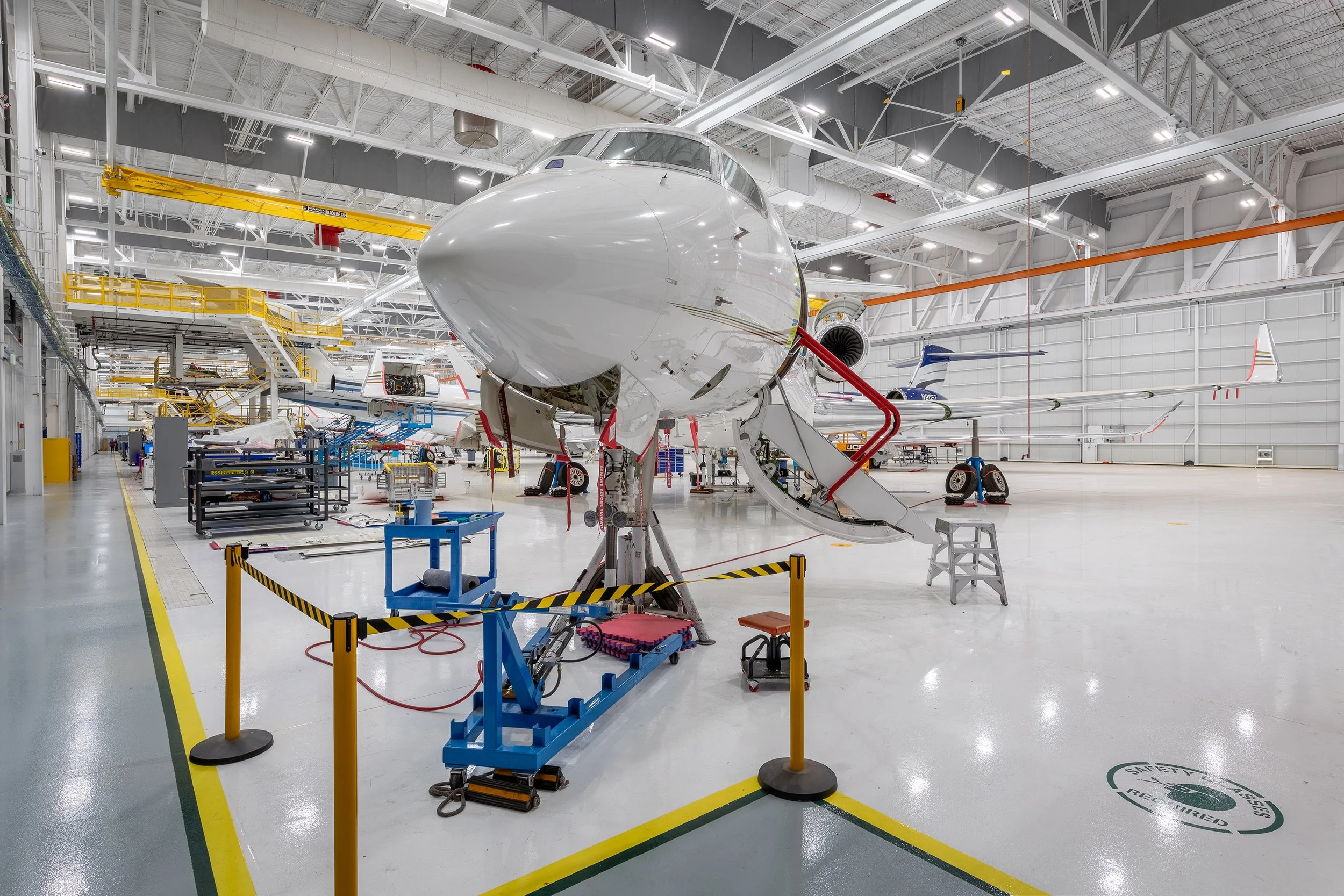 A large aircraft in a brightly lit, spacious hangar undergoing maintenance, surrounded by tools, equipment, and safety barriers. Other aircraft and aviation machinery are visible in the background, highlighting Arizona's bustling aerospace industry.