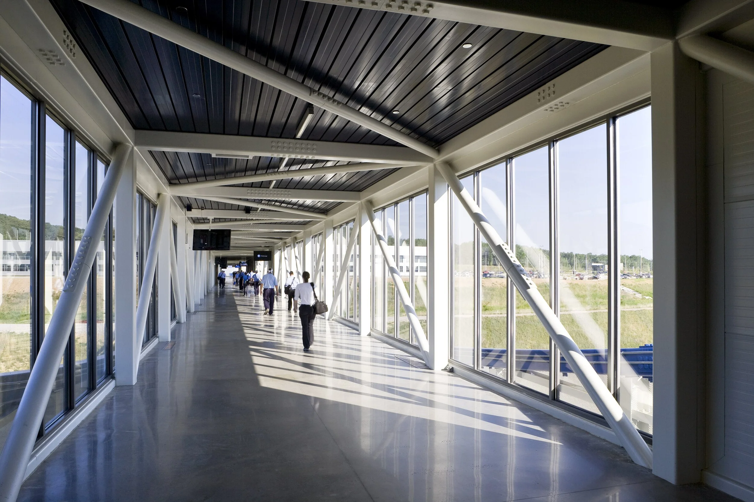 Employees walk through a bright, glass-walled Volkswagen pedestrian bridge with metal beams and a polished floor, modern industrial architecture.