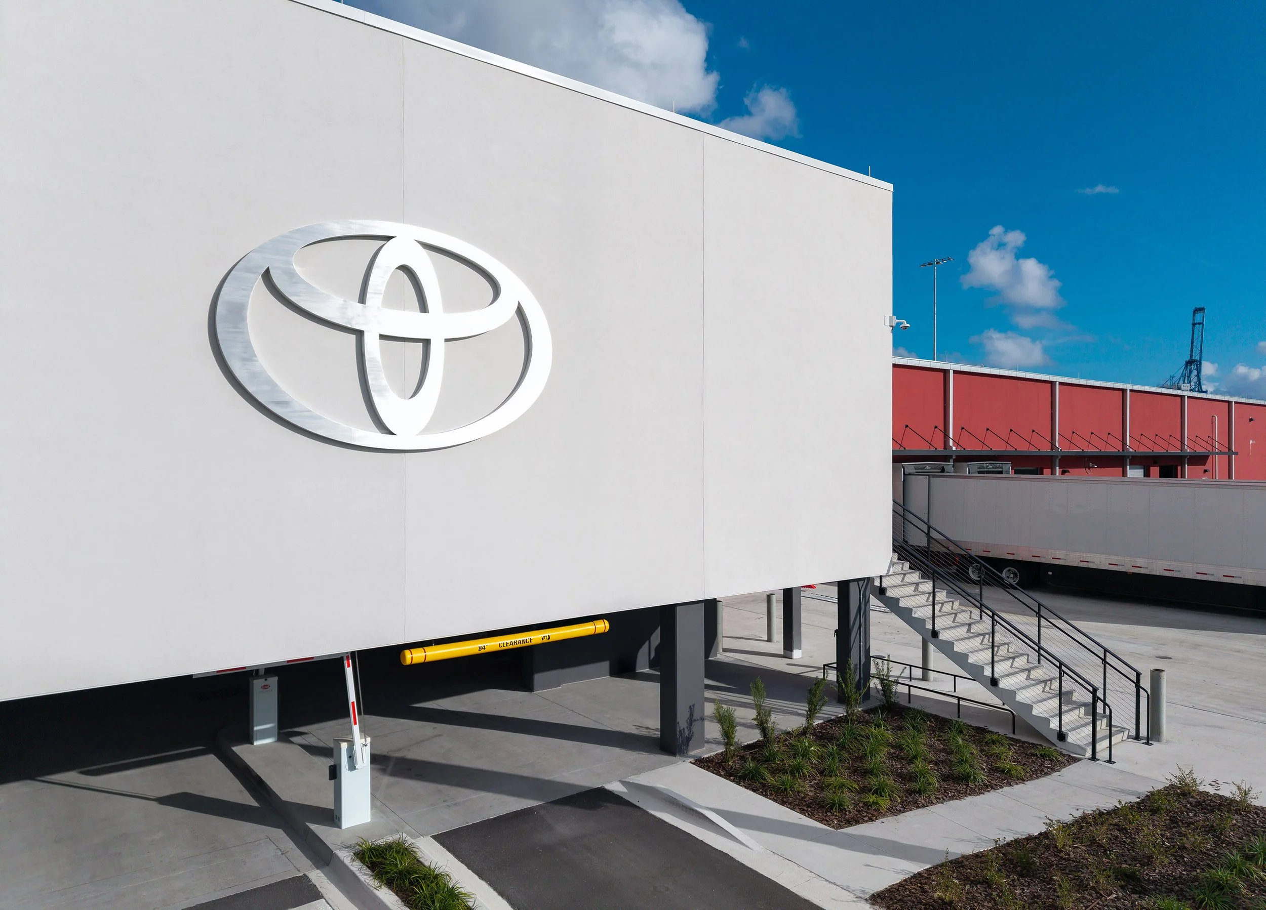 A large silver Toyota logo is mounted on the exterior wall of a modern industrial building near a loading dock, with a clear blue sky in the background and a semi-truck trailer parked alongside.