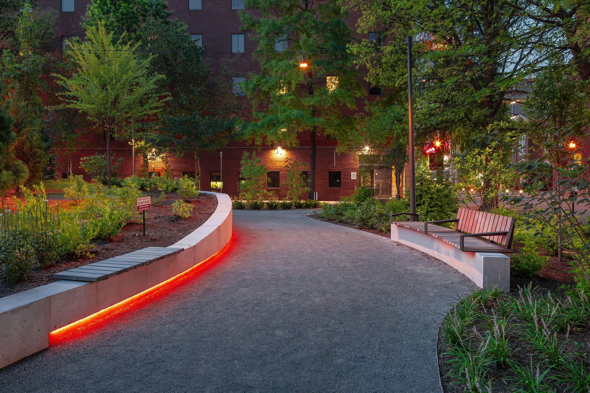 A curved urban pathway in a city park is illuminated by red LED lights along a low concrete wall, with benches on one side, lush greenery, and a brick building in the background at dusk, showcasing thoughtful landscape design.