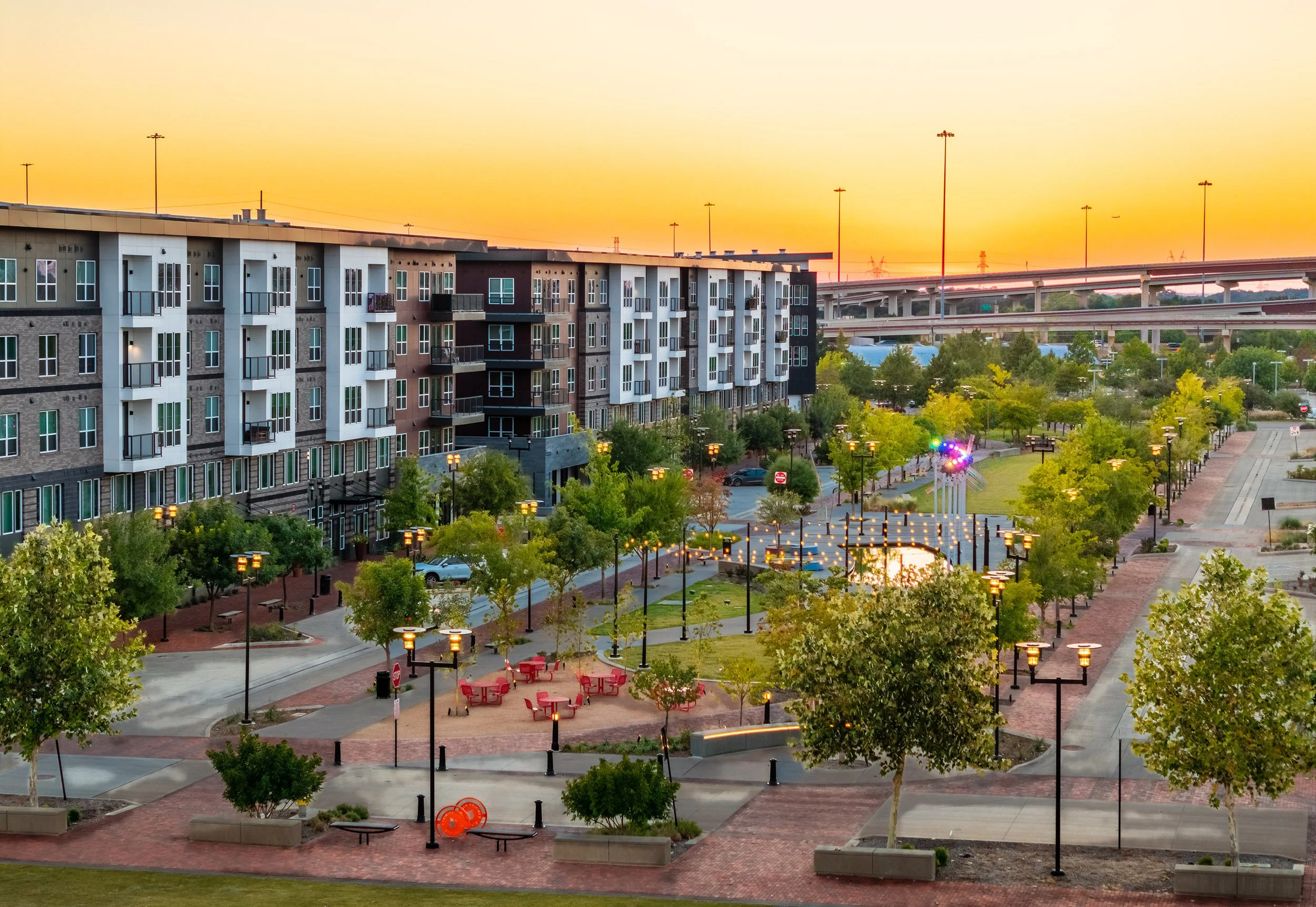A modern apartment complex overlooks a landscaped urban park with walking paths, trees, outdoor seating, and string lights at sunset. An elevated highway is visible in the background.