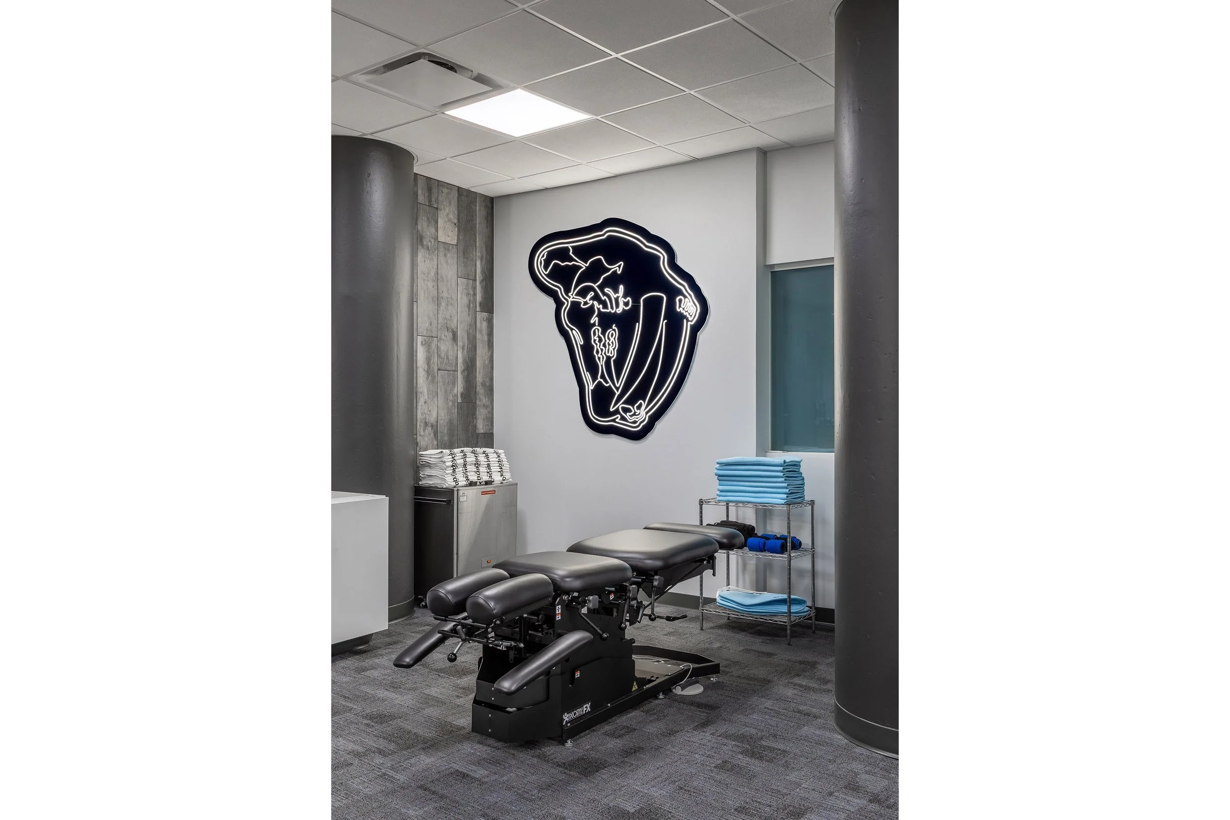 A modern chiropractic or physical therapy room designed for recovery, featuring a black adjustable treatment table, neatly stacked blue towels on a metal cart, and a large illuminated abstract wall art behind. Grey pillars and a tiled ceiling complet