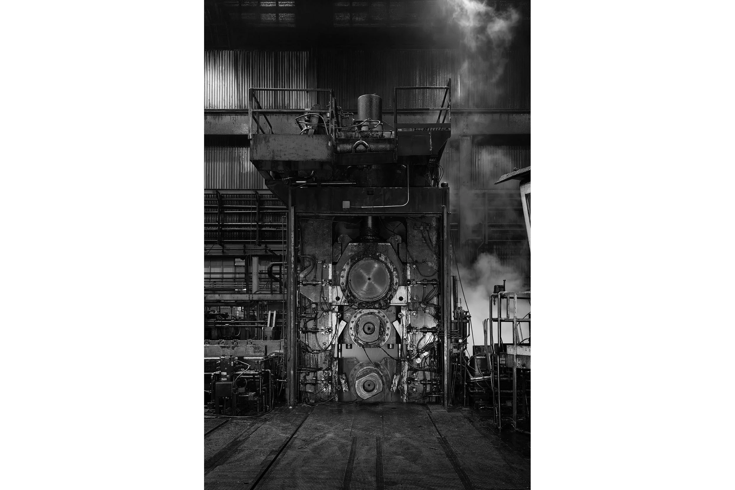 Large aluminum industrial machine in a factory setting, surrounded by pipes and steam, in black and white.