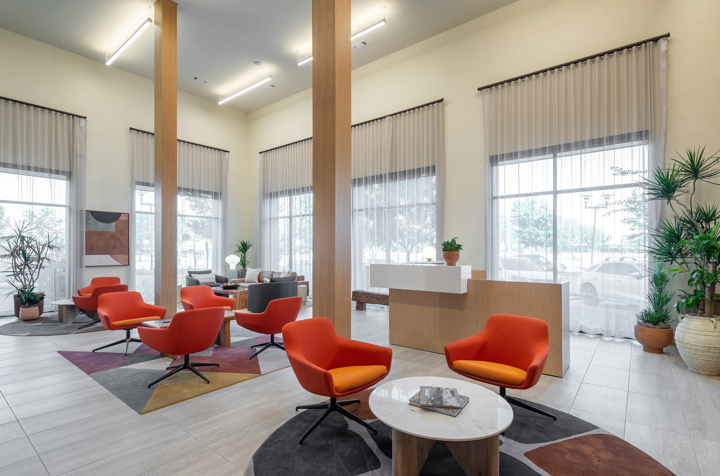 Modern lobby with tall windows, sheer curtains, light flooring, and wooden columns. Features orange chairs around small tables, a reception desk, indoor plants, and a bright, welcoming atmosphere.