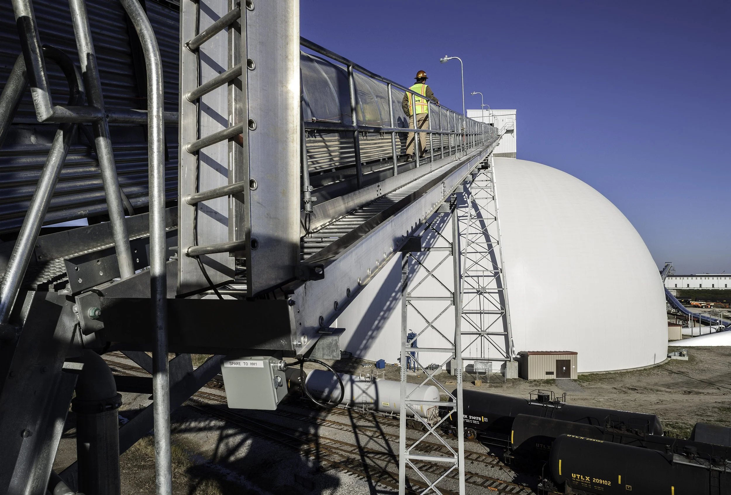 A worker walks on an elevated metal walkway near a large white dome and train tracks under a clear blue sky.