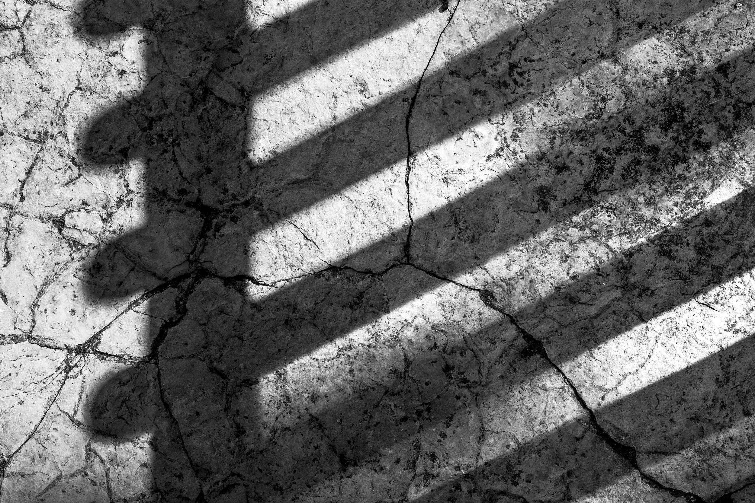Diagonal shadows of a fence cast on cracked stone walk, evoking a winter stillness near a Shaker building.