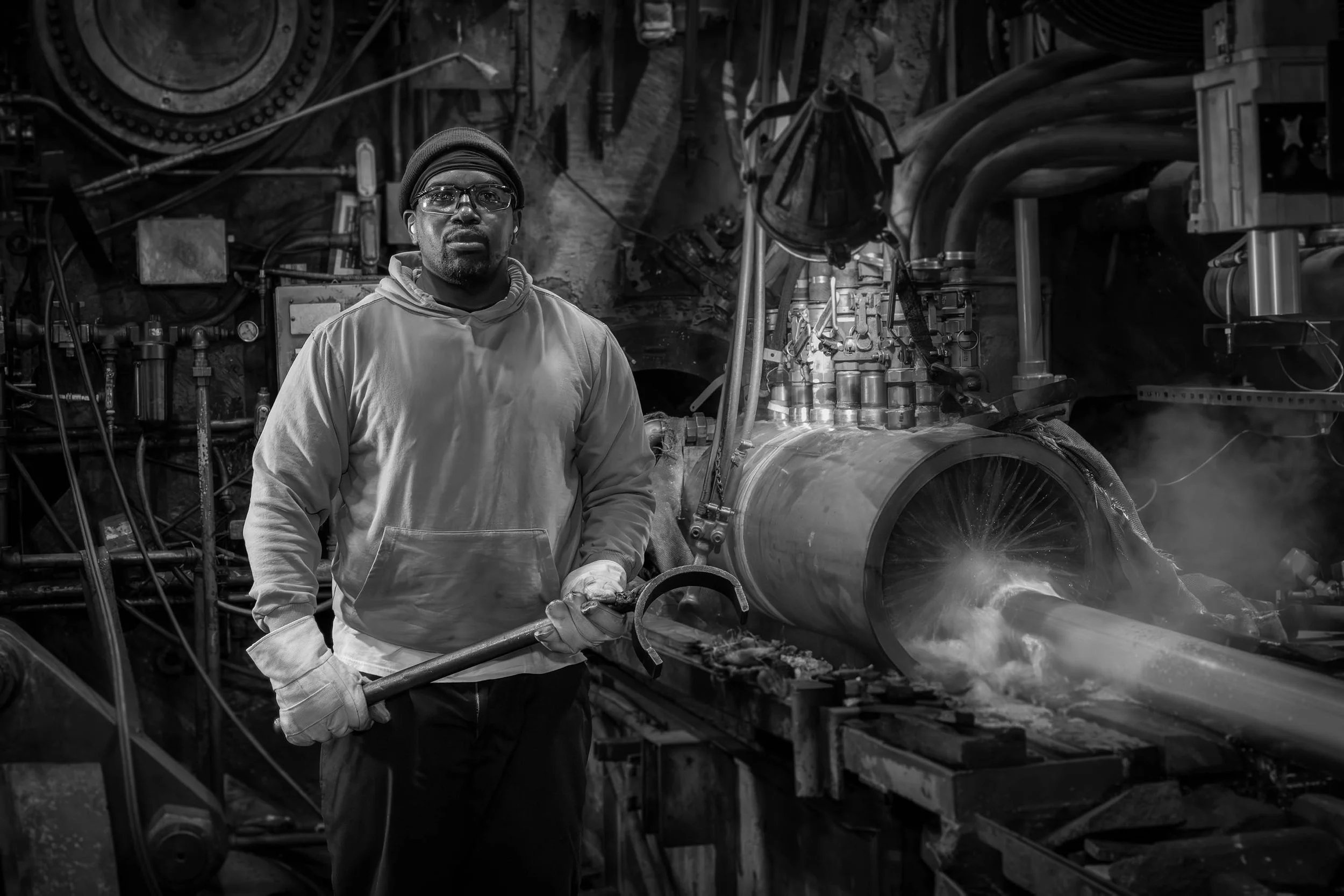 A man in work clothes stands in a factory, embodying the industrial workforce as he holds a large tool beside steam-emitting machinery.