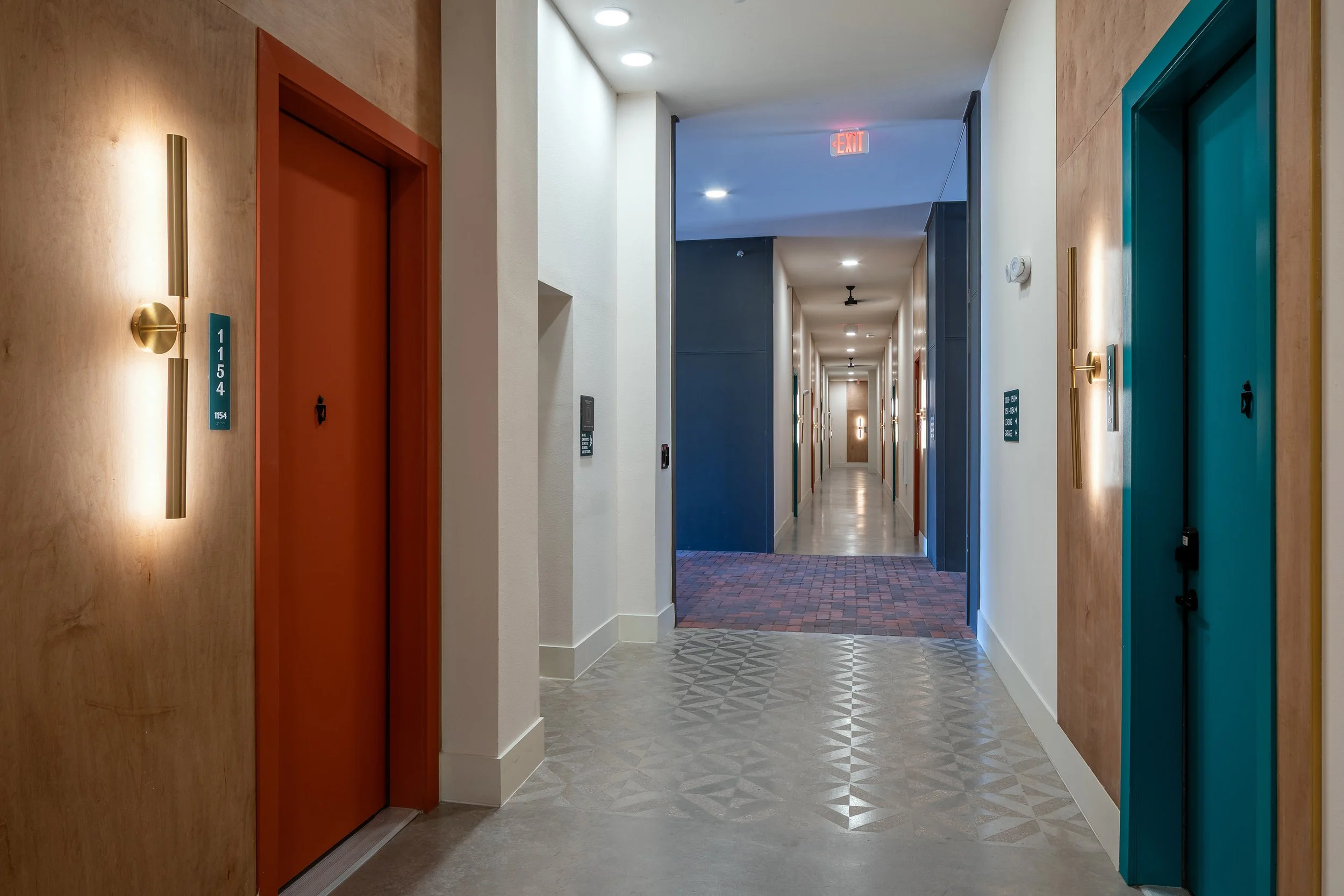 Modern hallway with patterned tile floor, wood-paneled walls, and two elevators—one with a red door and another with a teal door. Bright lighting and a long corridor extend into the distance.