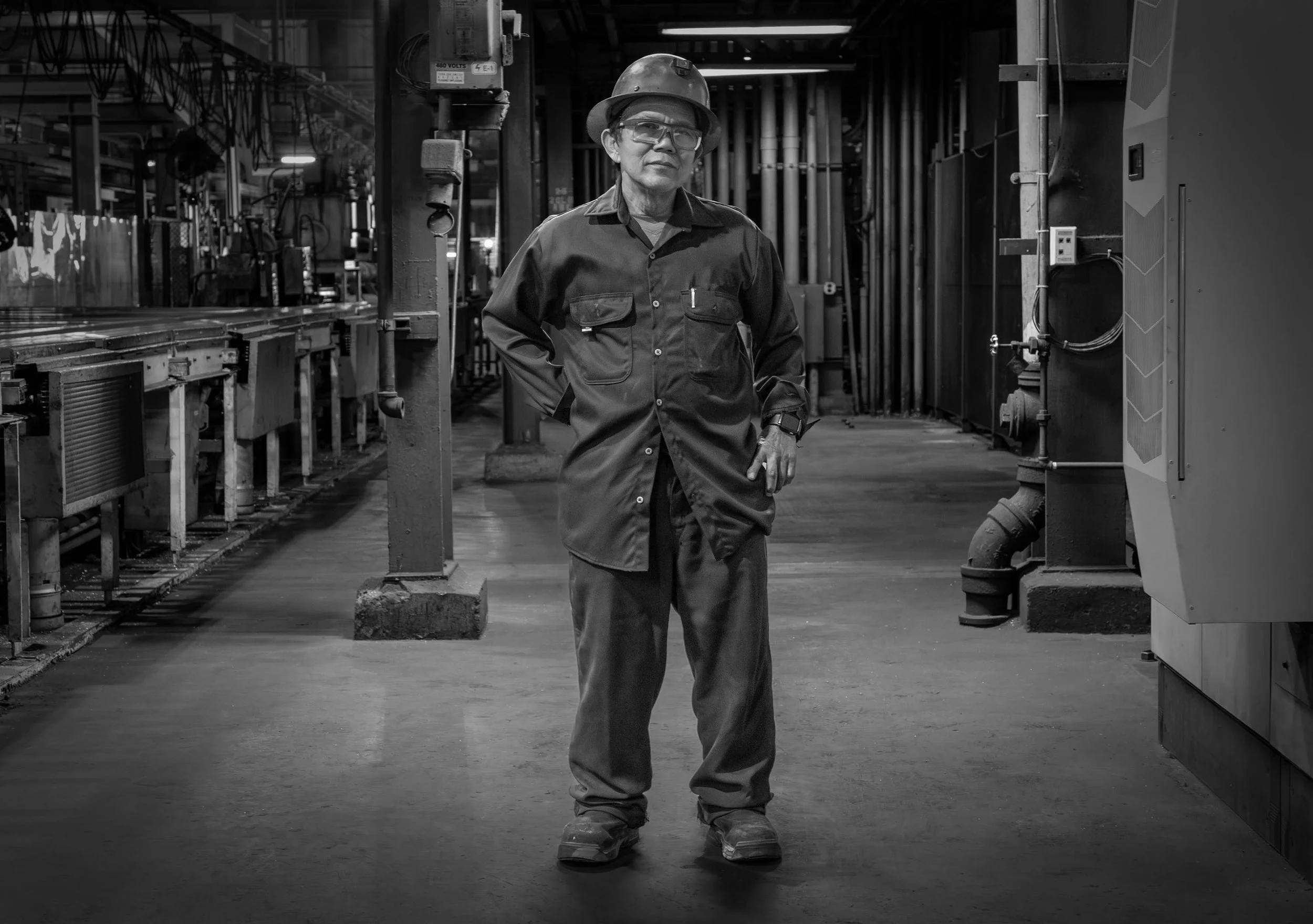 A factory worker in uniform and helmet stands amid aluminum stacks in a black and white industrial warehouse, facing the camera.