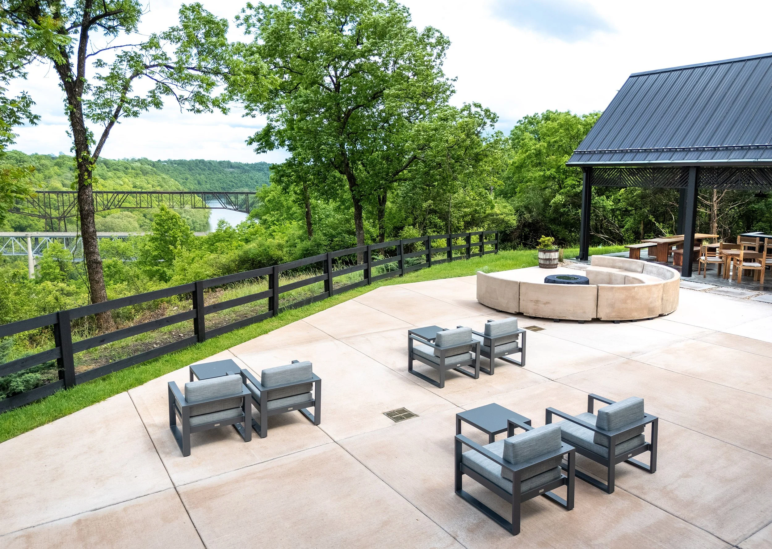 A spacious outdoor patio with modern gray chairs arranged around a circular fire pit, overlooking a lush green landscape and river. Unique architecture complements the covered seating area and wooden fence under a partly cloudy sky.