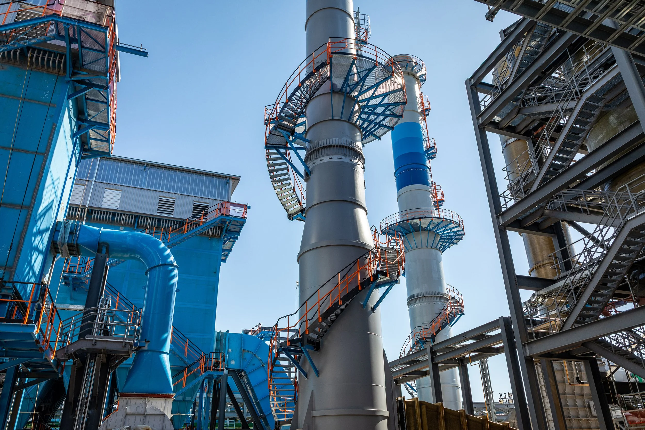 A modern industrial facility features large blue and gray metal structures, architectural cylindrical towers with spiral staircases, and an intricate process of pipes and platforms under a clear sky.