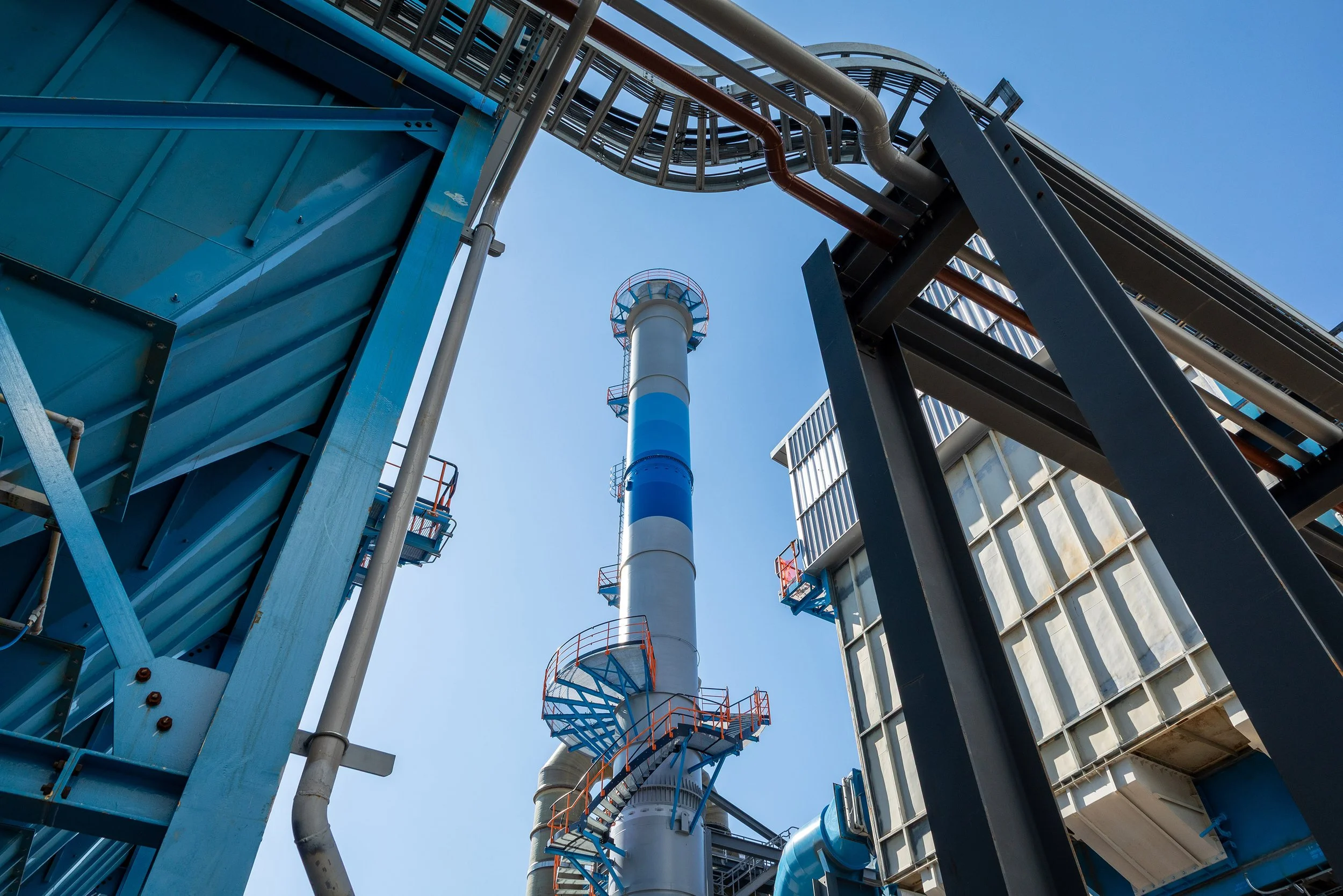 A view looking up at an industrial facility with a tall, cylindrical smokestack featuring blue stripes, surrounded by architectural metal structures, staircases, and walkways against a clear blue sky.