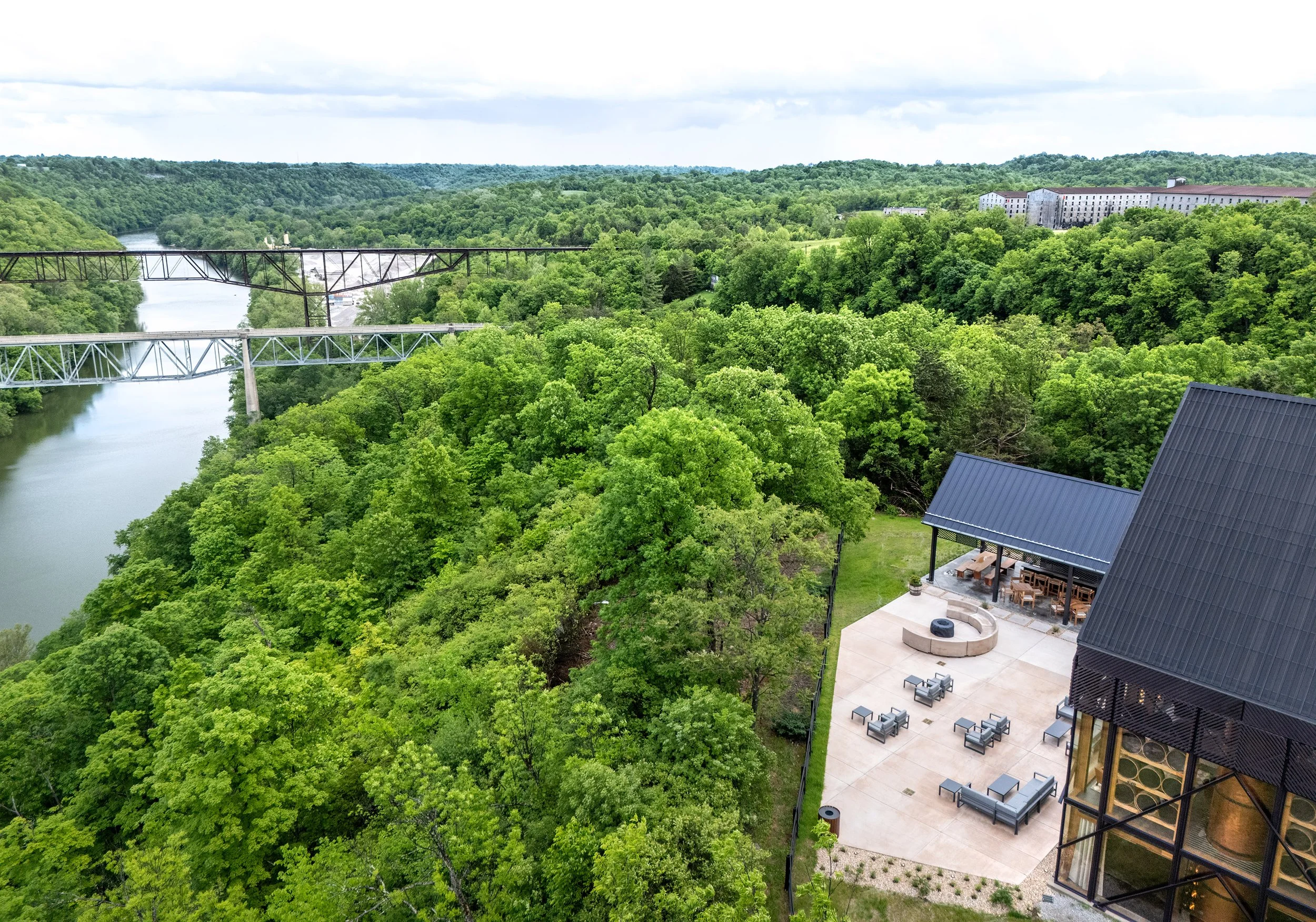 A modern visitor center with striking architecture and outdoor seating is nestled among lush green trees beside a river, with a bridge and rolling hills visible in the background under a cloudy sky.