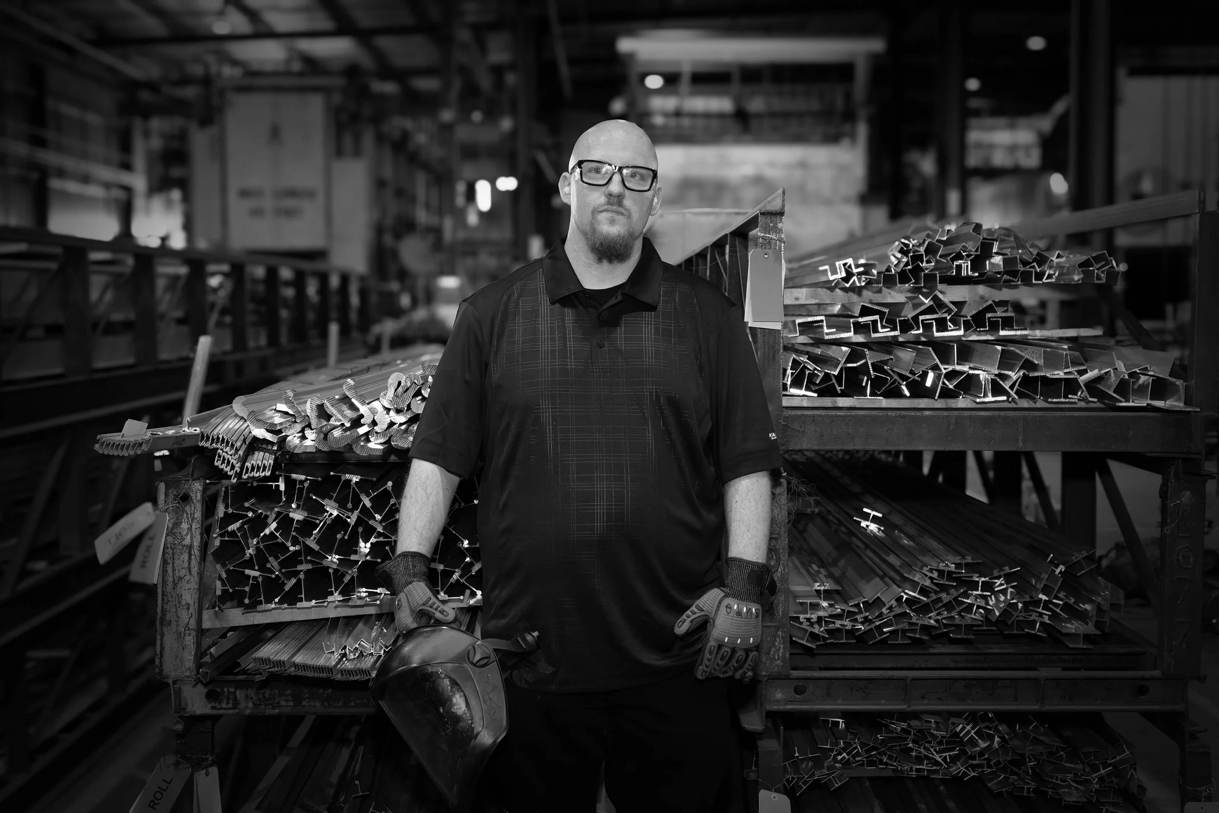 A man in work gloves and glasses stands in a factory beside aluminum rods, holding a welding mask.