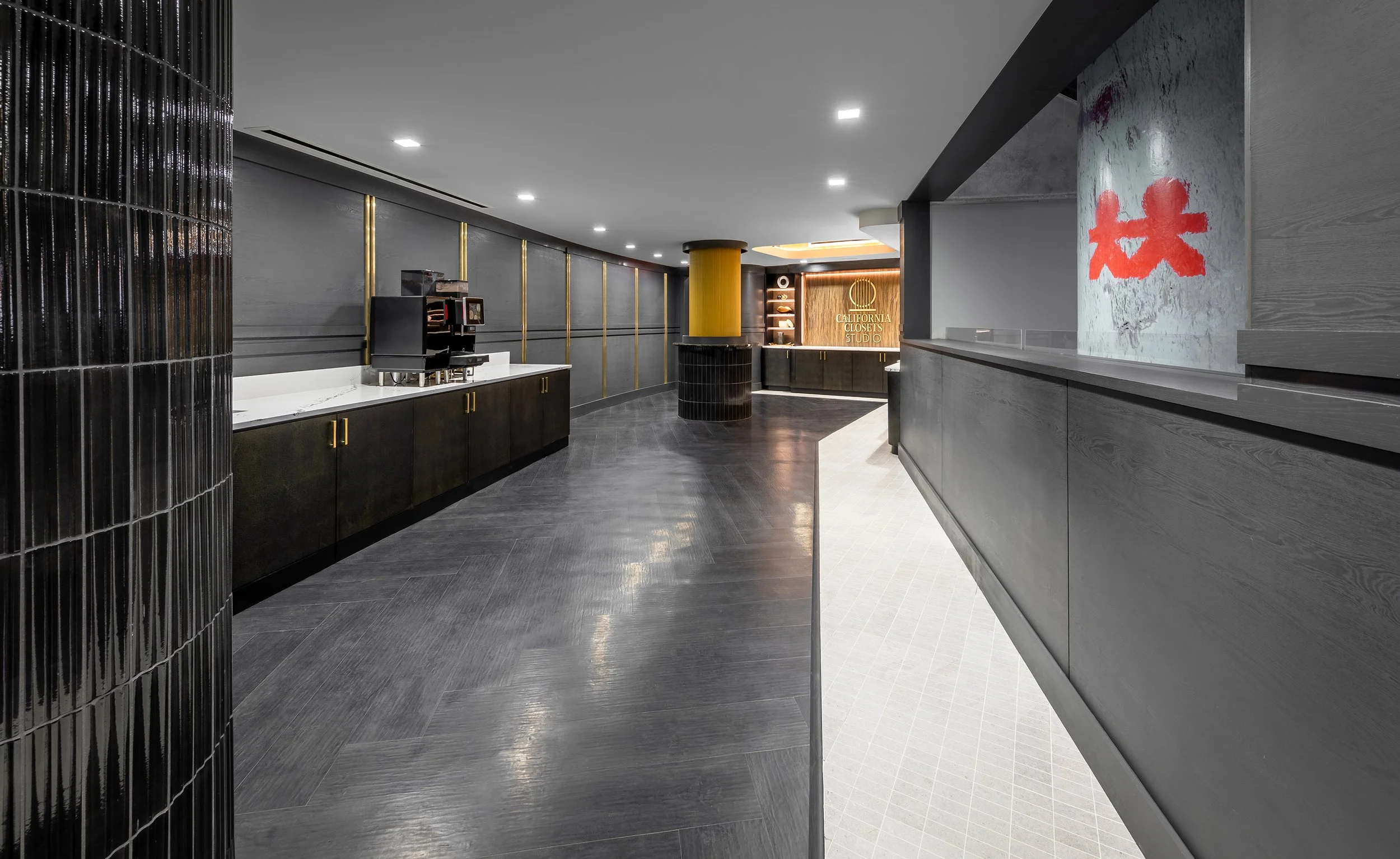 A modern, sleek suite lobby with dark flooring, black cabinets, a coffee machine, wood accents, recessed lighting, and a glass wall featuring red painted marks. The space curves towards a warmly lit luxury food service area.