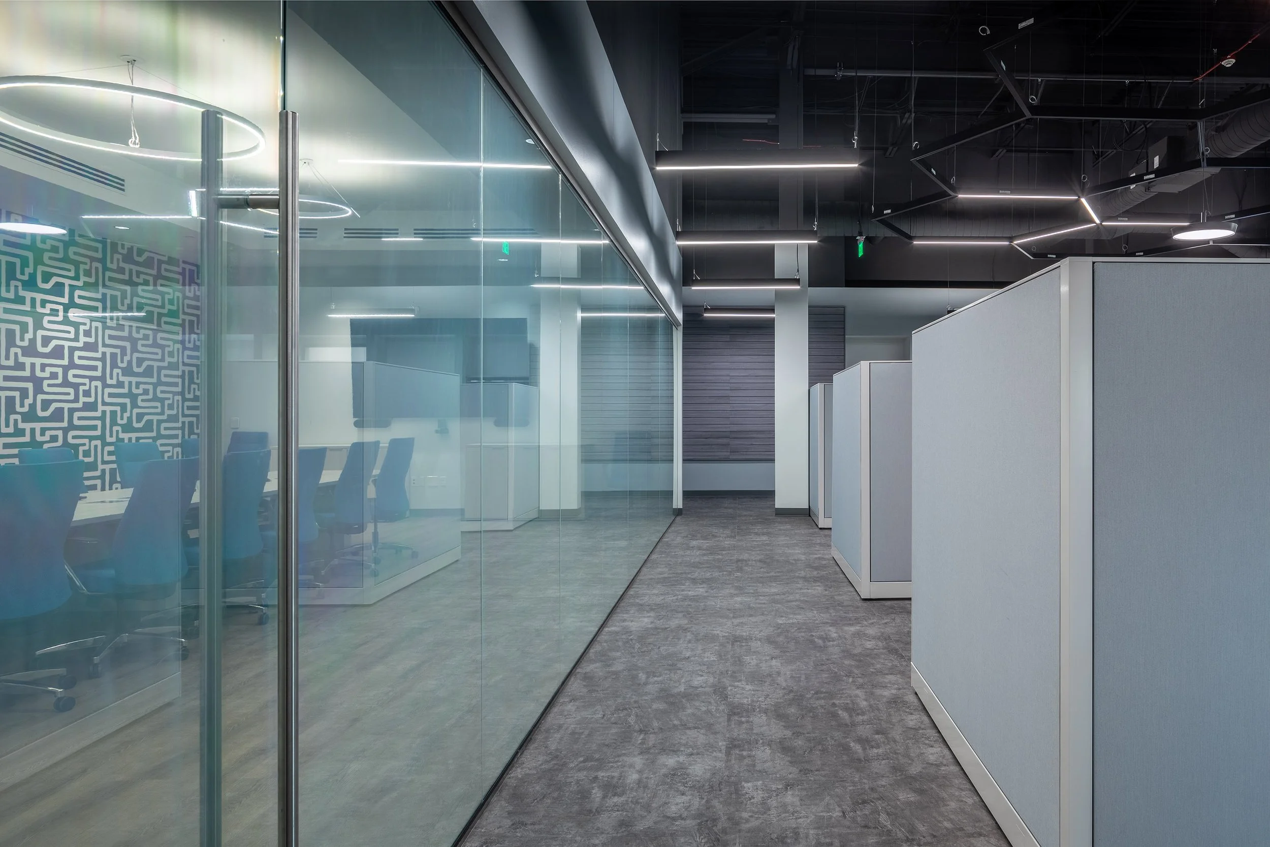 Modern office space with glass-walled conference room on the left and cubicles with light blue panels on the right.