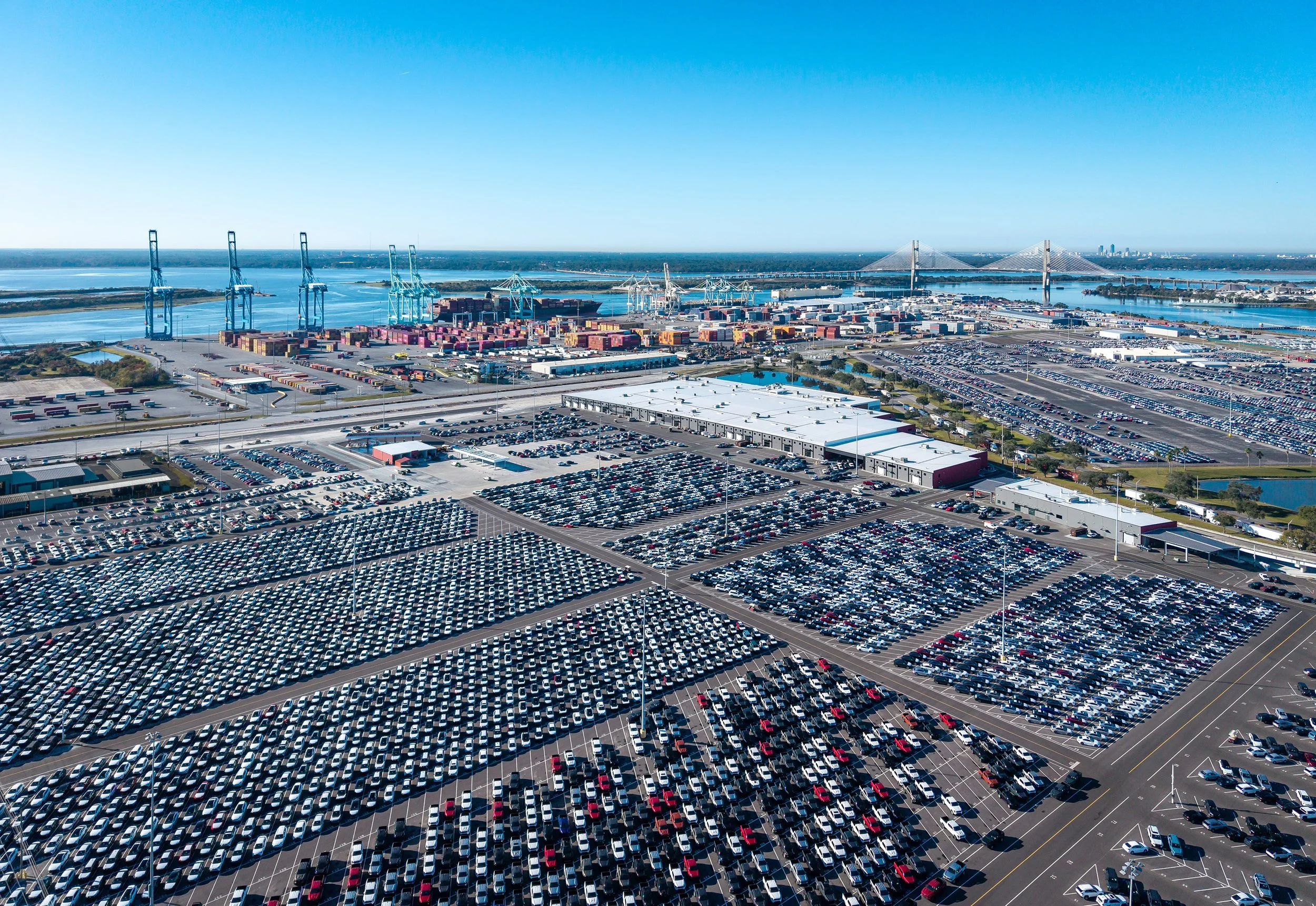 Aerial view of a large port with thousands of parked cars, shipping containers, cranes, and a distant suspension bridge under a clear blue sky near a body of water.