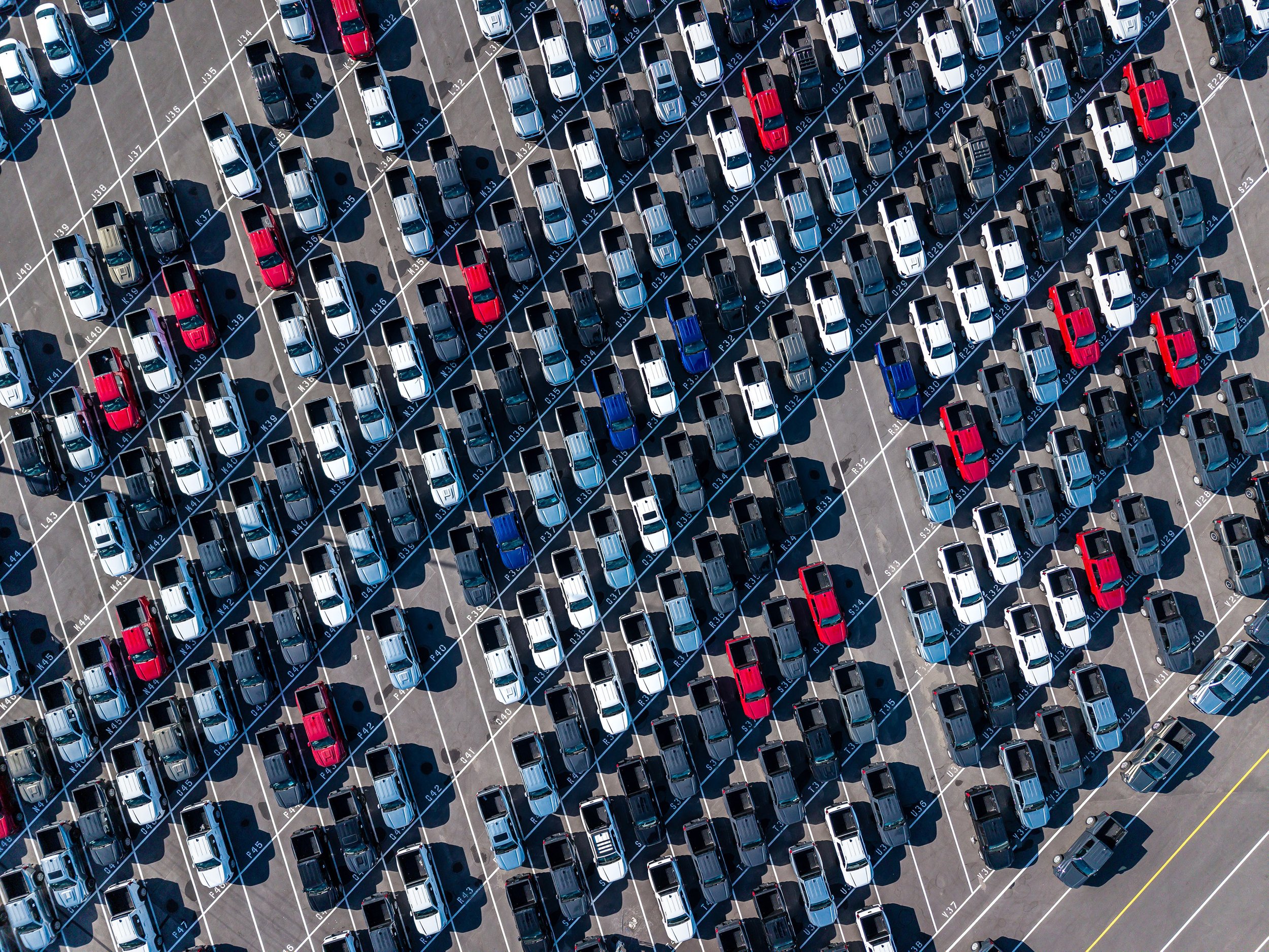 Aerial view of a large parking lot filled with rows of parked cars in various colors, including white, black, red, and blue, organized in neat, diagonal lines.