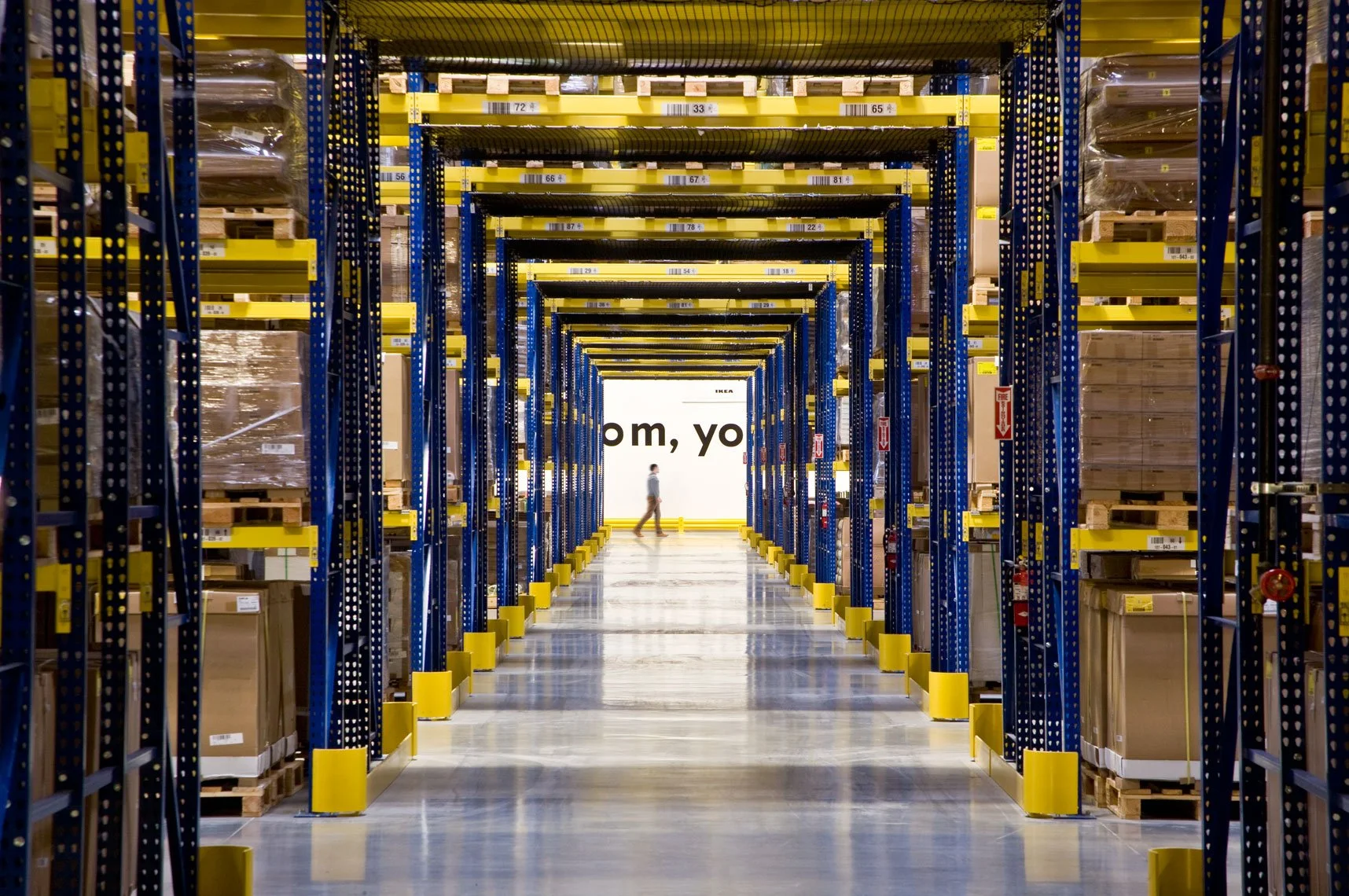 View of a warehouse aisle with tall shelves stocked with boxes, leading to a distant wall with partially visible text.