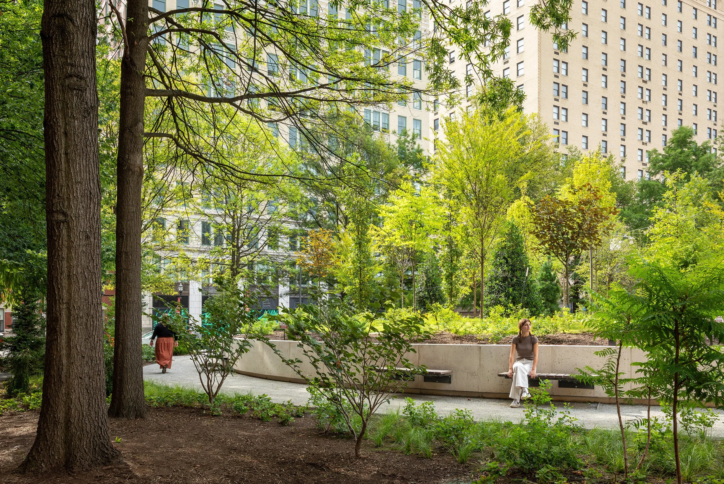 A woman sits on a curved bench in an urban park landscape surrounded by trees and plants, with tall buildings visible in the background on a sunny day. Another person walks along a path nearby.